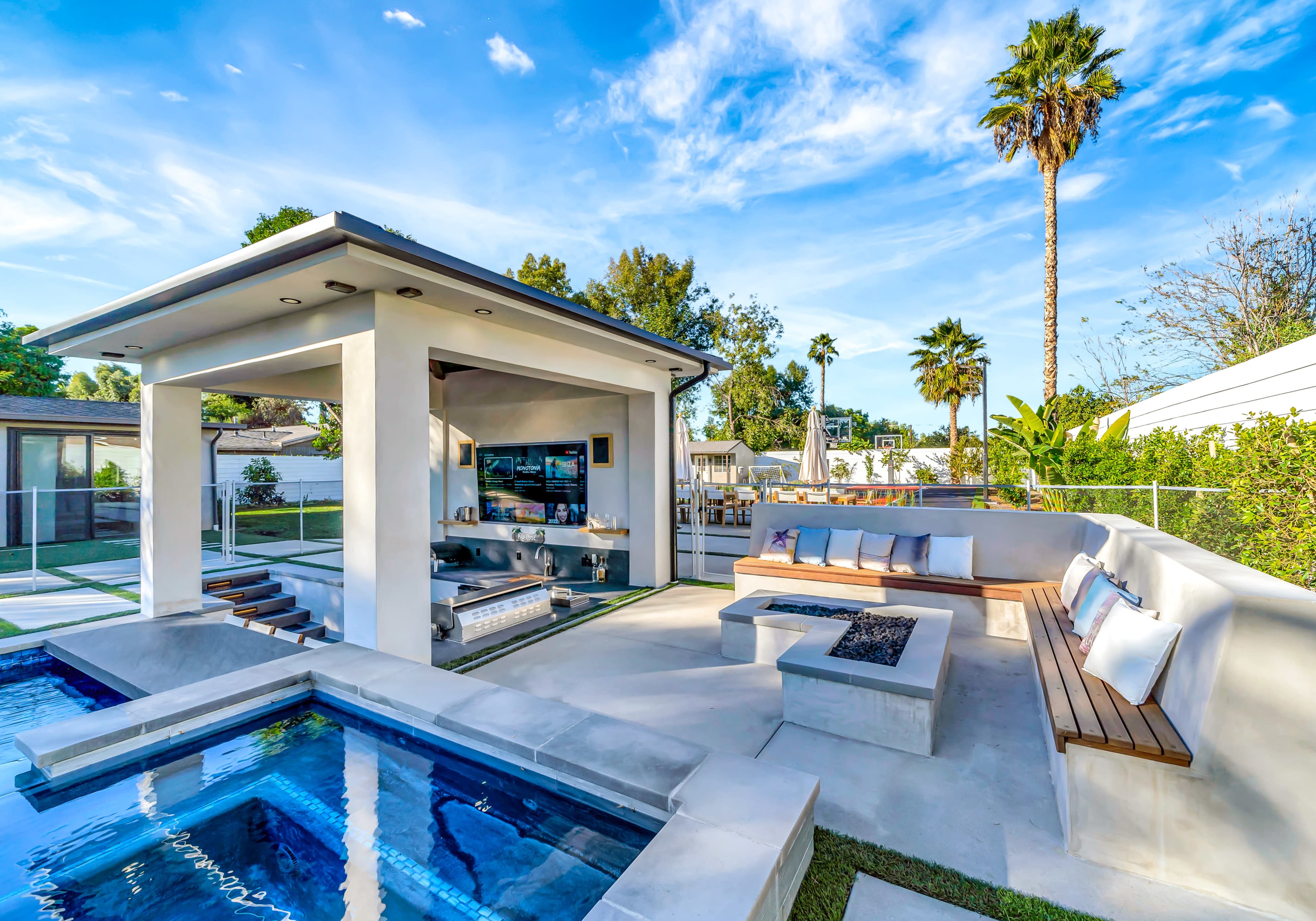 A modern outdoor lounge area with a pool, a covered seating space, and palm trees in the background.