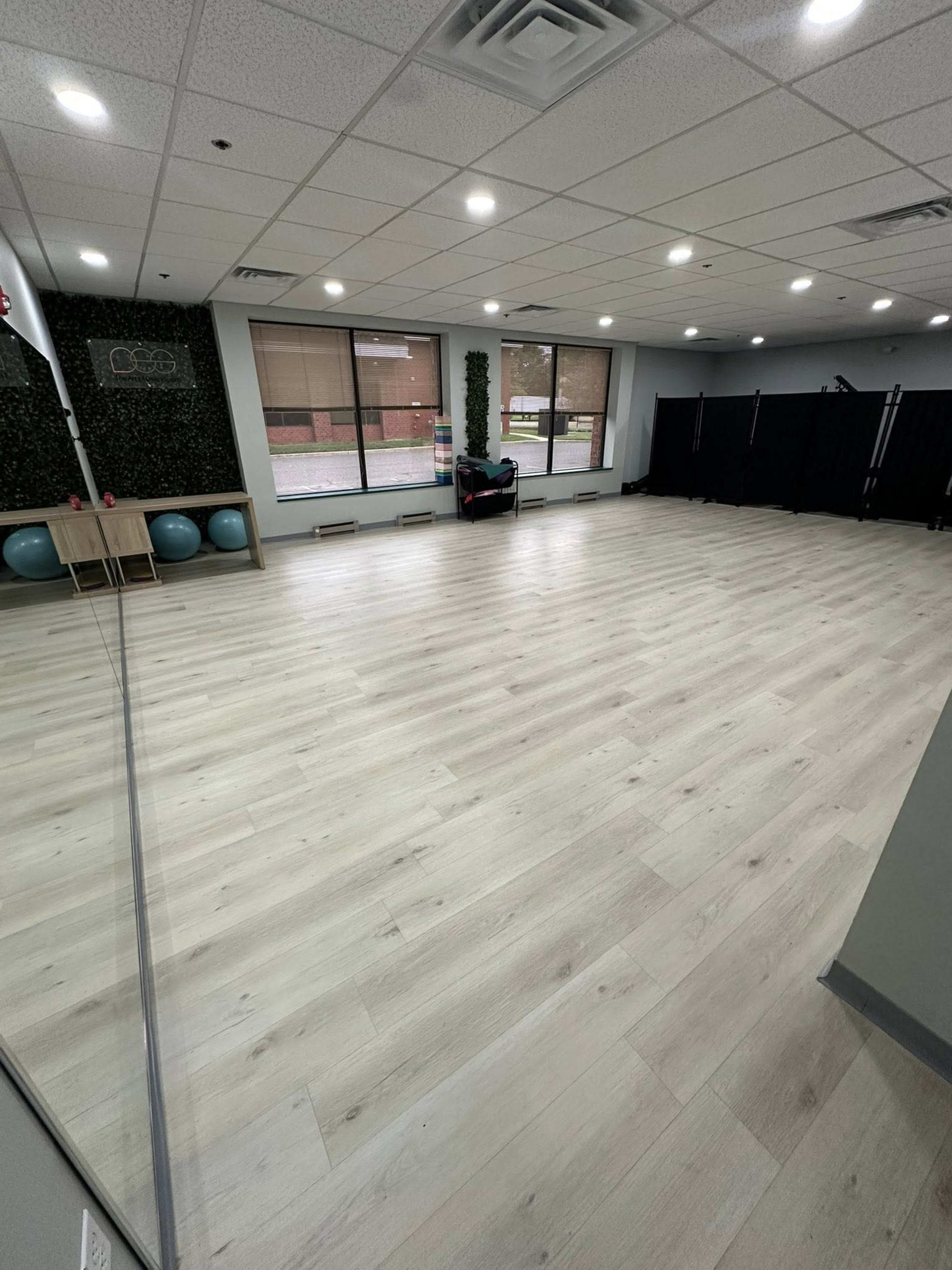 The image shows a spacious studio with wooden flooring, large windows, and fitness equipment in the corner.