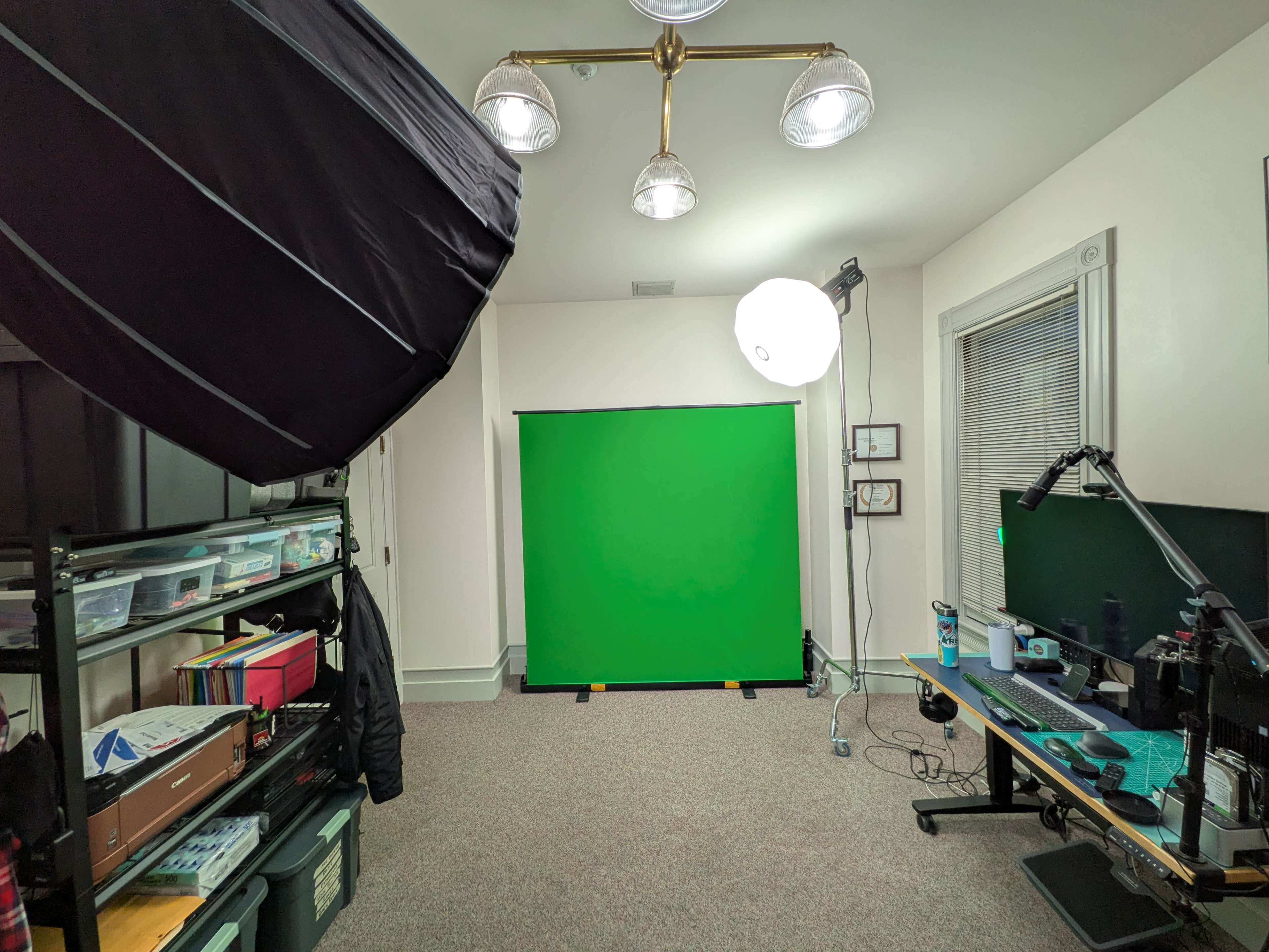 The image shows a room set up for video production, featuring a green screen backdrop, lighting equipment, and a desk with a monitor.