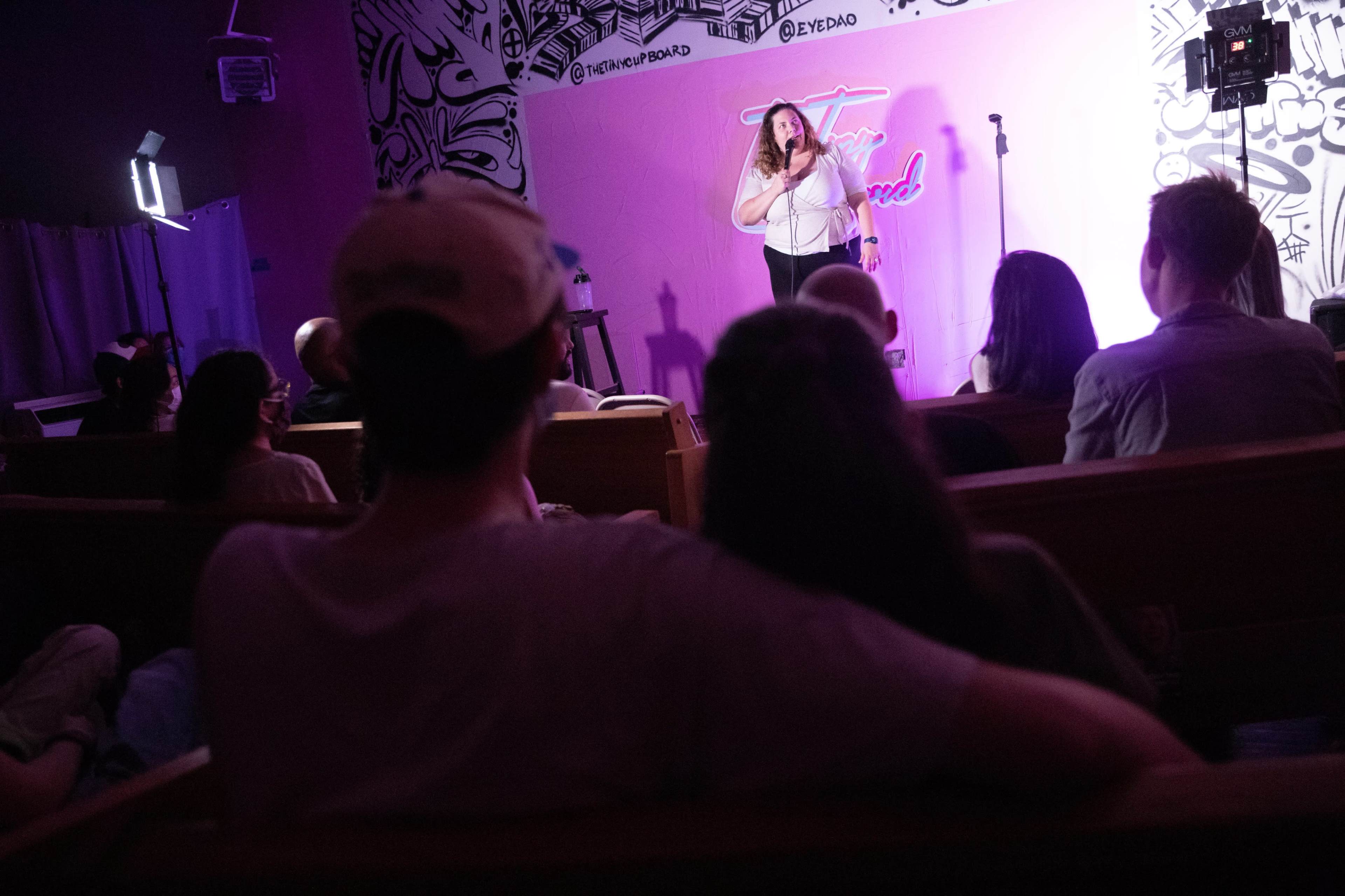 A performer stands on stage speaking into a microphone while an audience watches from wooden benches in a brightly decorated venue.