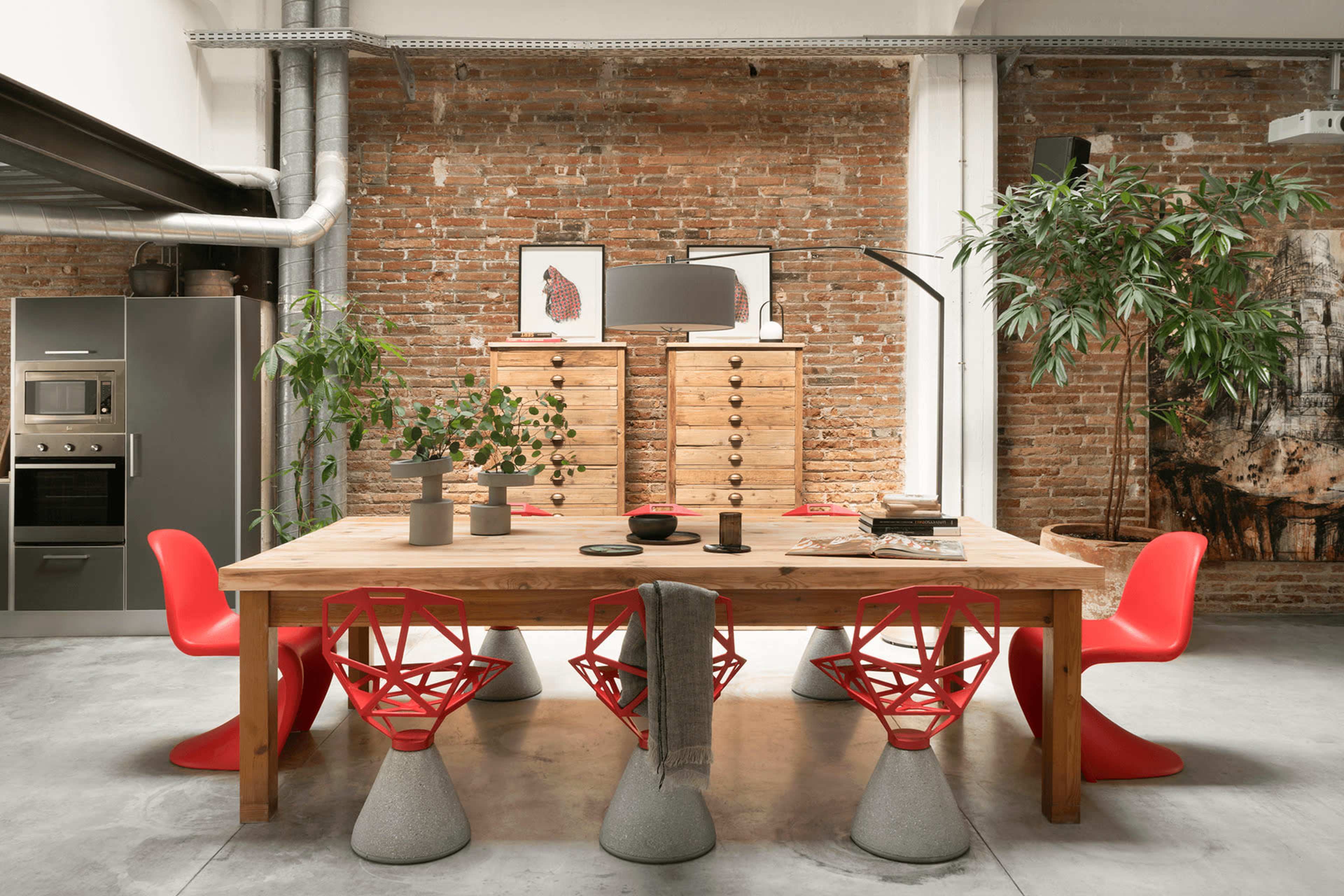 The image shows a modern dining area featuring a wooden table surrounded by red chairs, with exposed brick walls and a minimalist kitchen in the background.