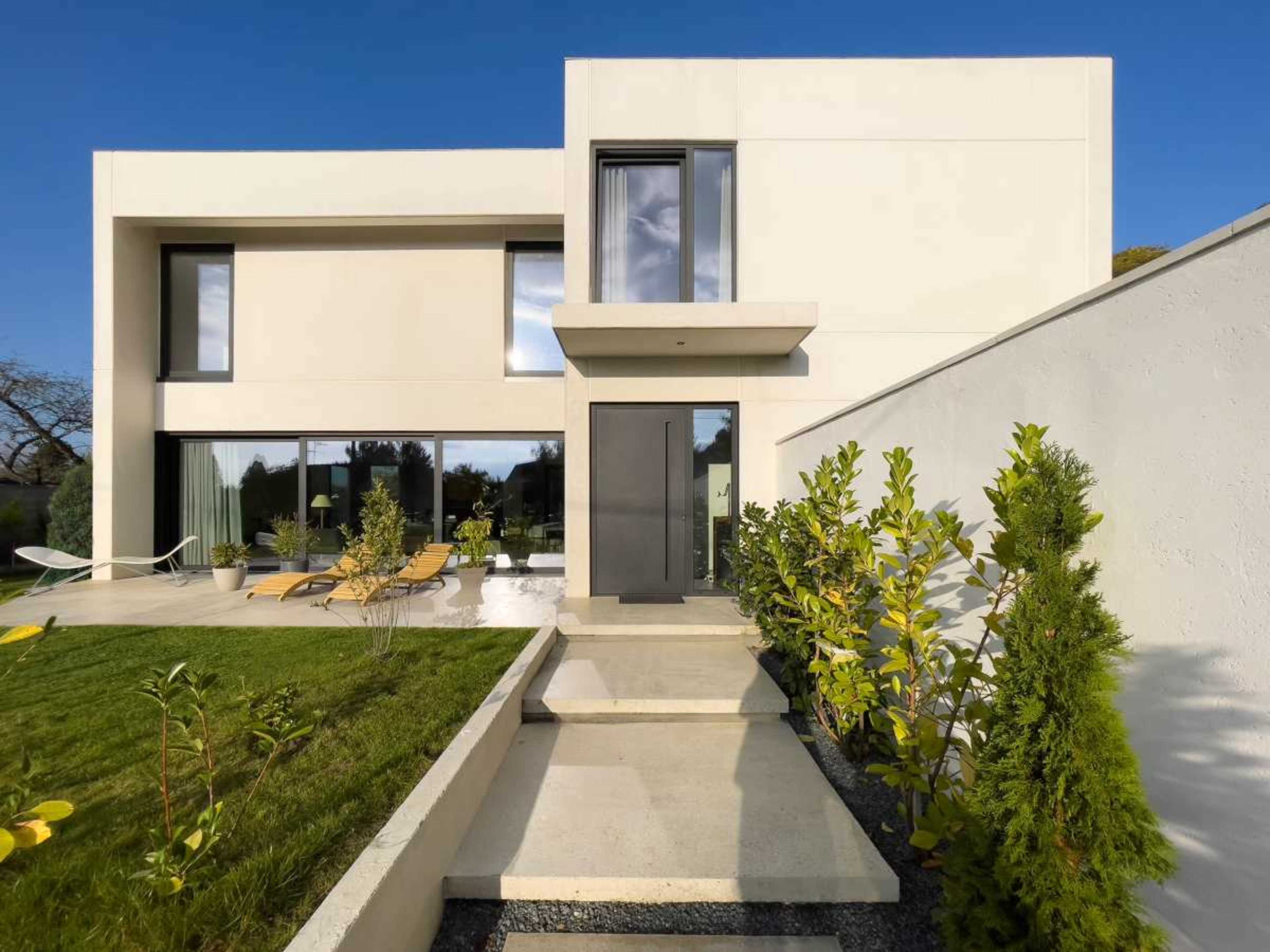 The image shows a modern two-story house with a minimalist design, featuring large windows, a black front door, and a landscaped garden with plants and a small pathway leading to the entrance.