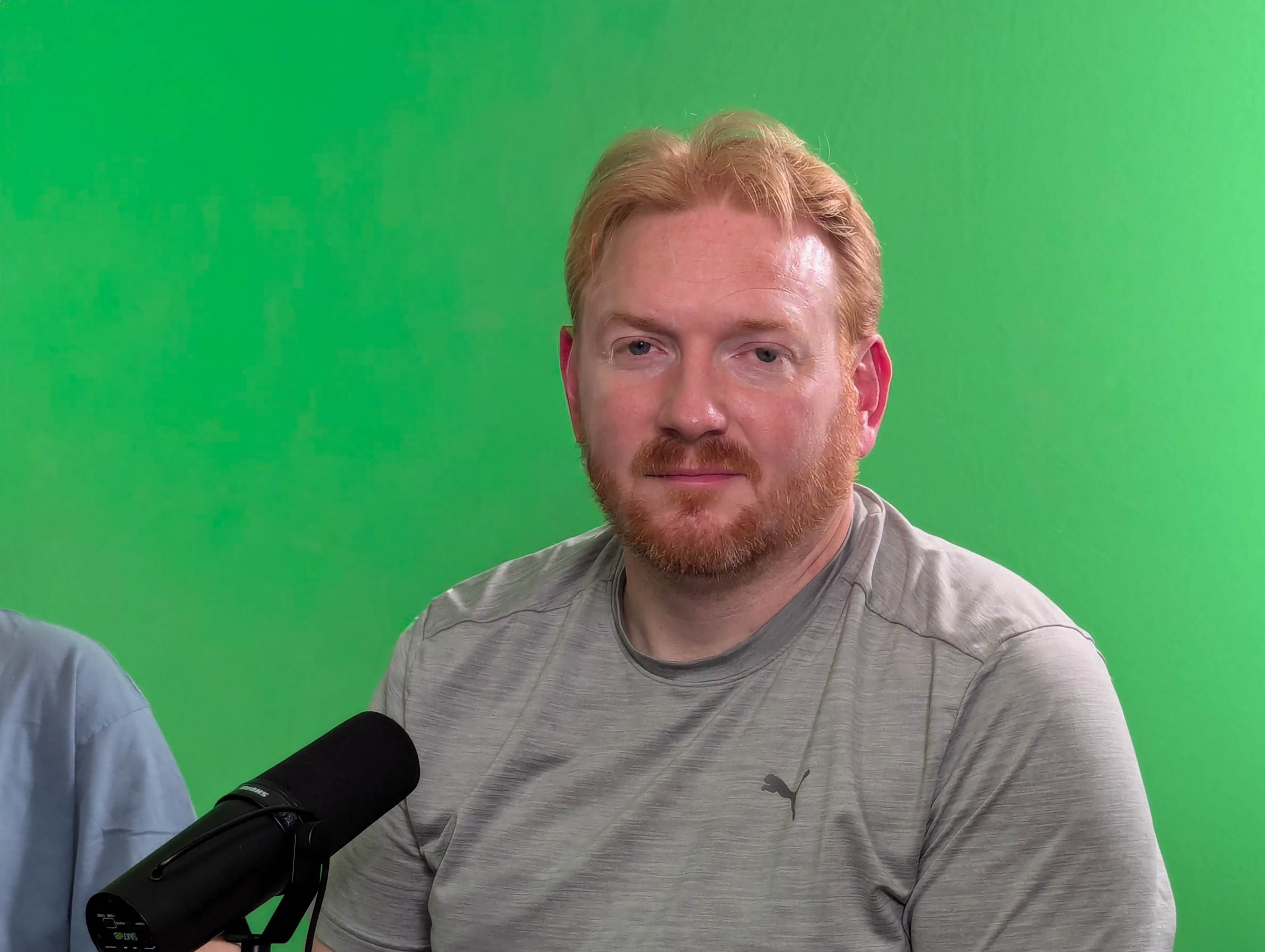 A man with a beard is sitting in front of a green screen, next to a microphone.