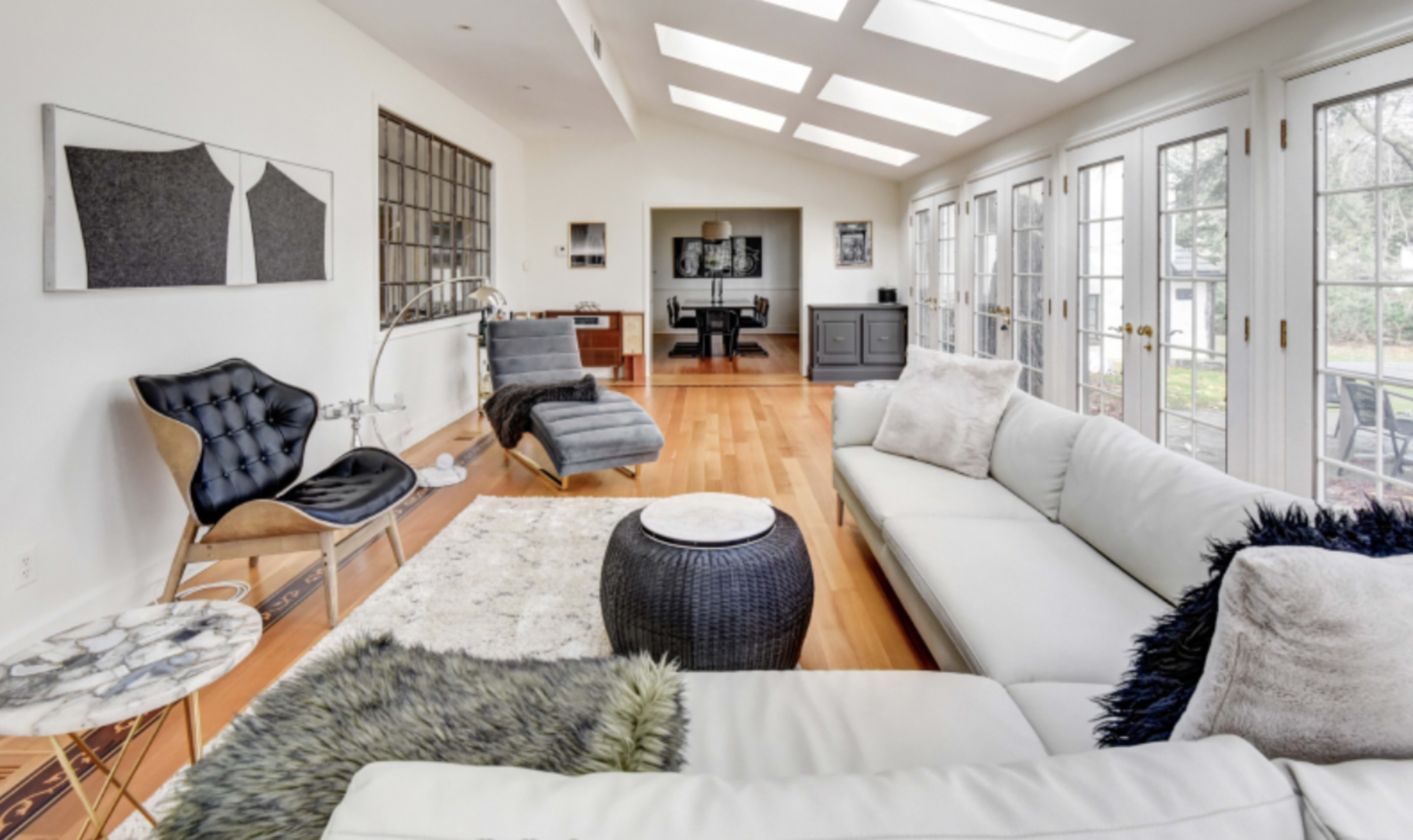 A modern, bright living space featuring large windows, a white sectional sofa, and various pieces of stylish furniture arranged around a wooden floor.