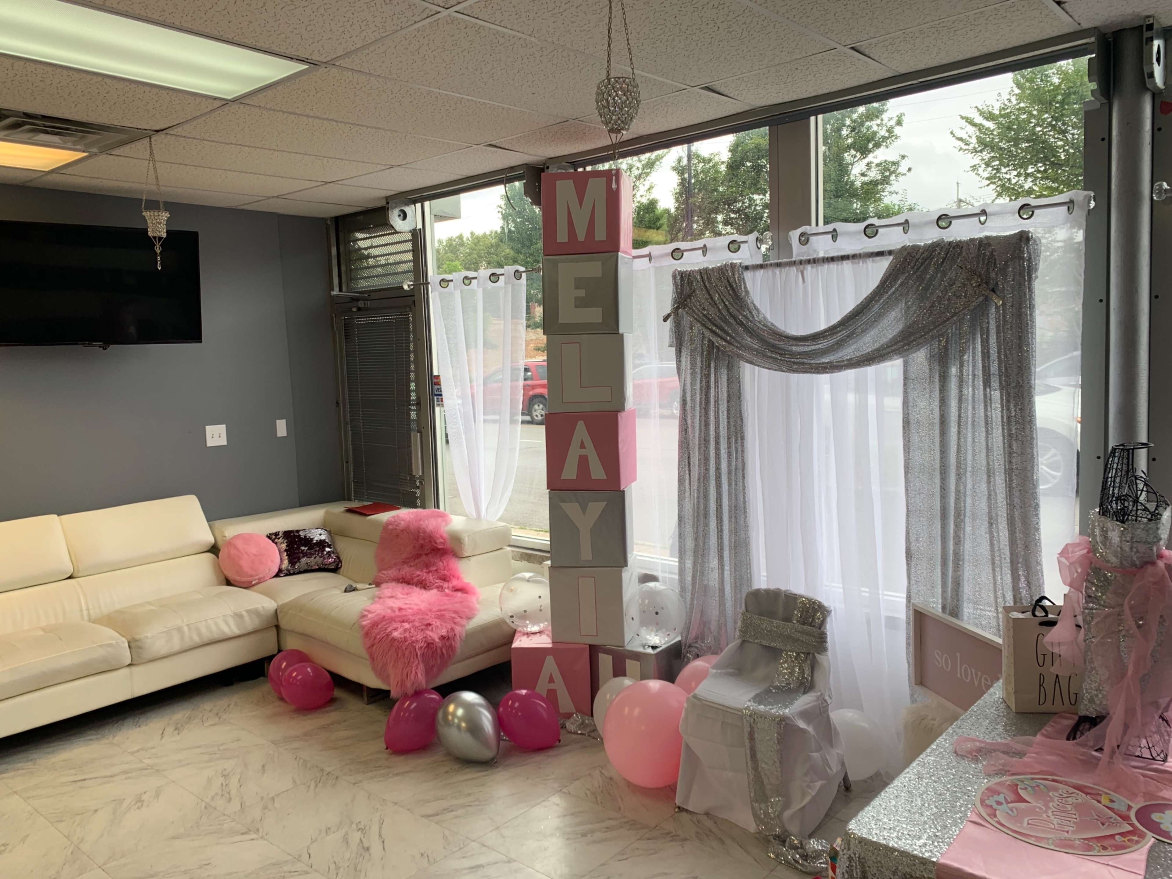 A decorated event space featuring a white couch, pink and silver balloons, and a large sign spelling "MELAYIA" against a backdrop of sheer curtains.