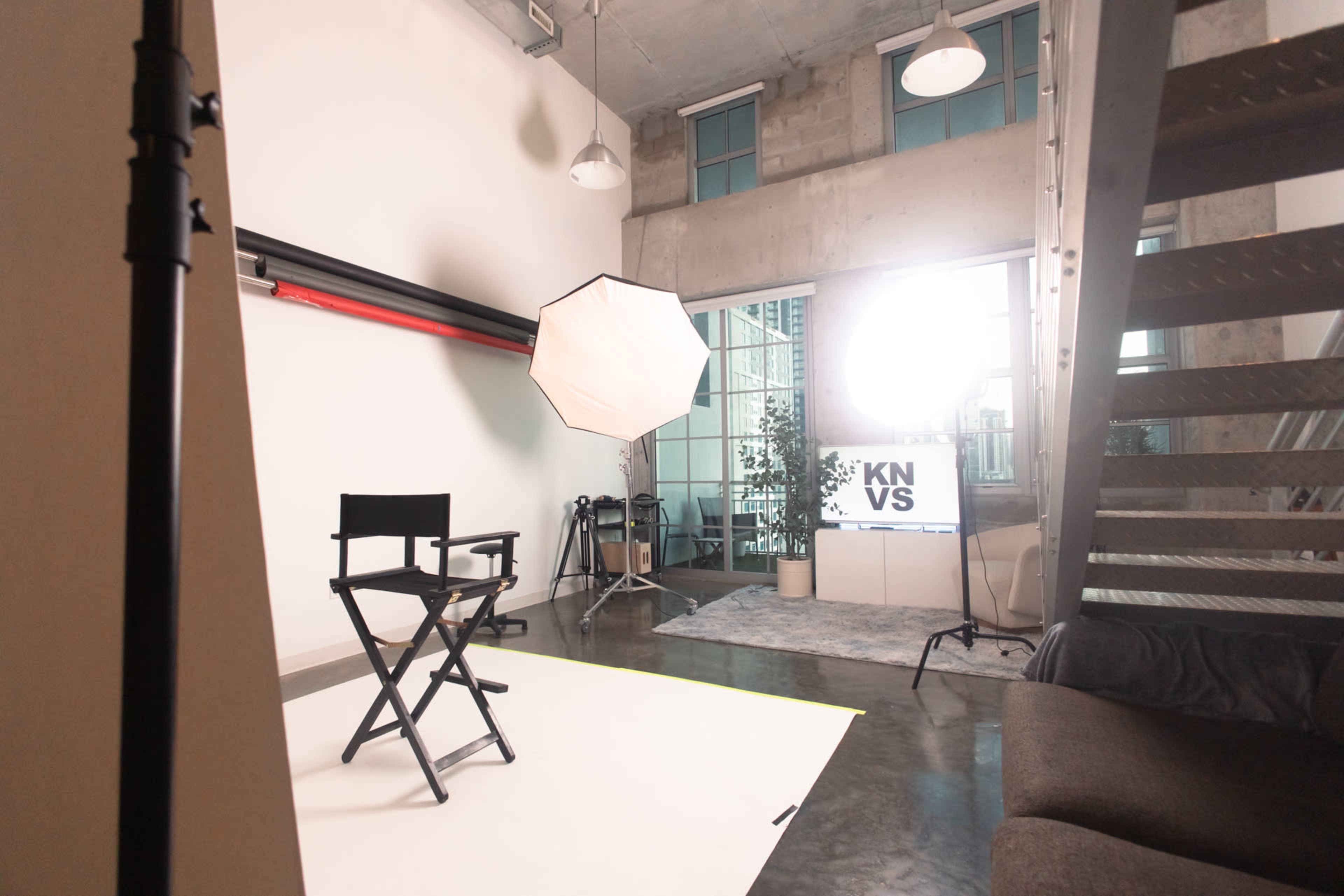 The image shows a photography studio setting with a director's chair, a large softbox light, and a backdrop, along with a monitor displaying text, in a well-lit industrial space.