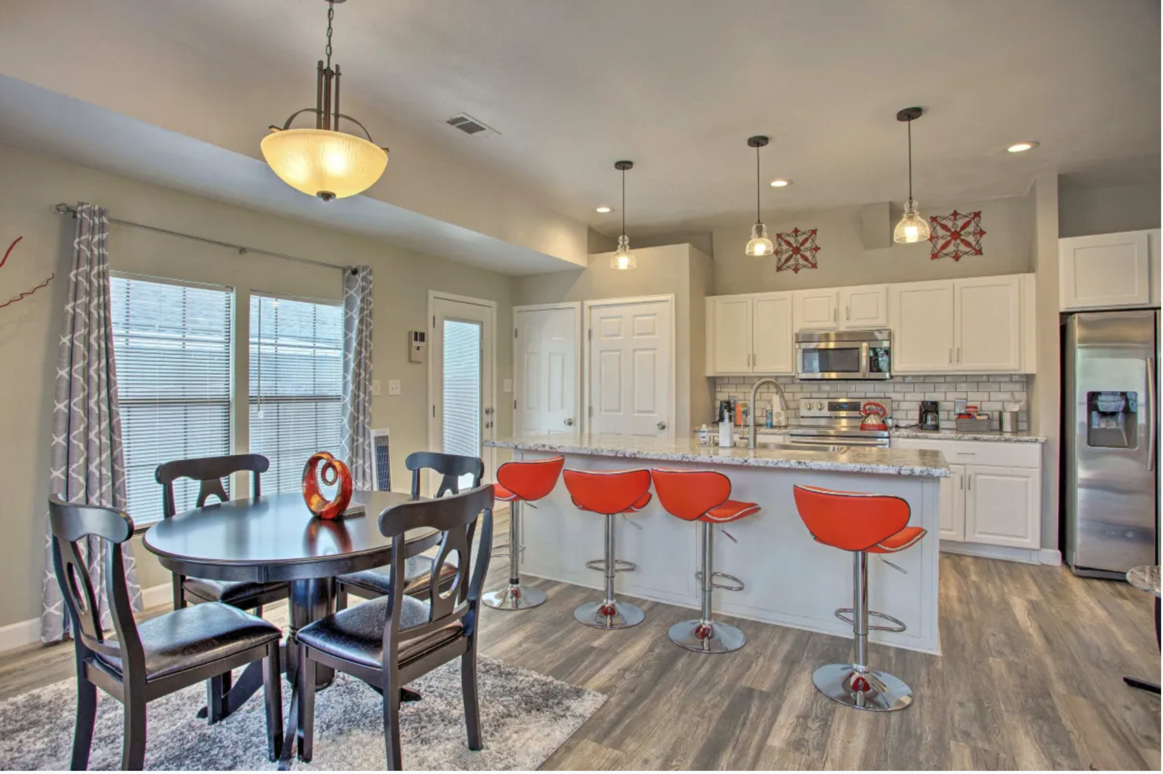 A modern kitchen with a dining area, featuring a round table with black chairs and red bar stools at a kitchen island, all set within a brightly lit space.