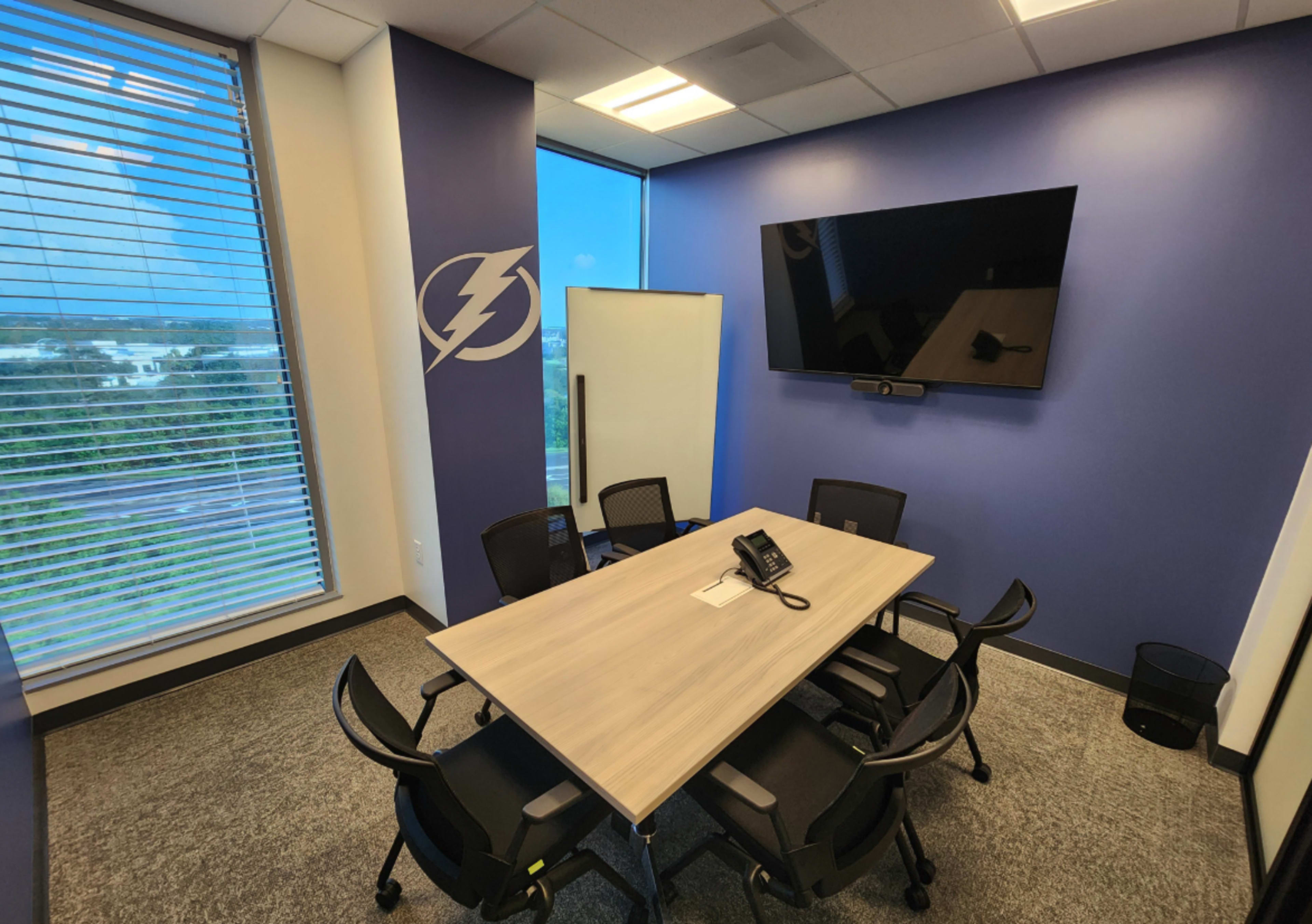 The image shows a small conference room with a rectangular table surrounded by six chairs, equipped with a wall-mounted TV and a whiteboard, set against a backdrop of large windows.