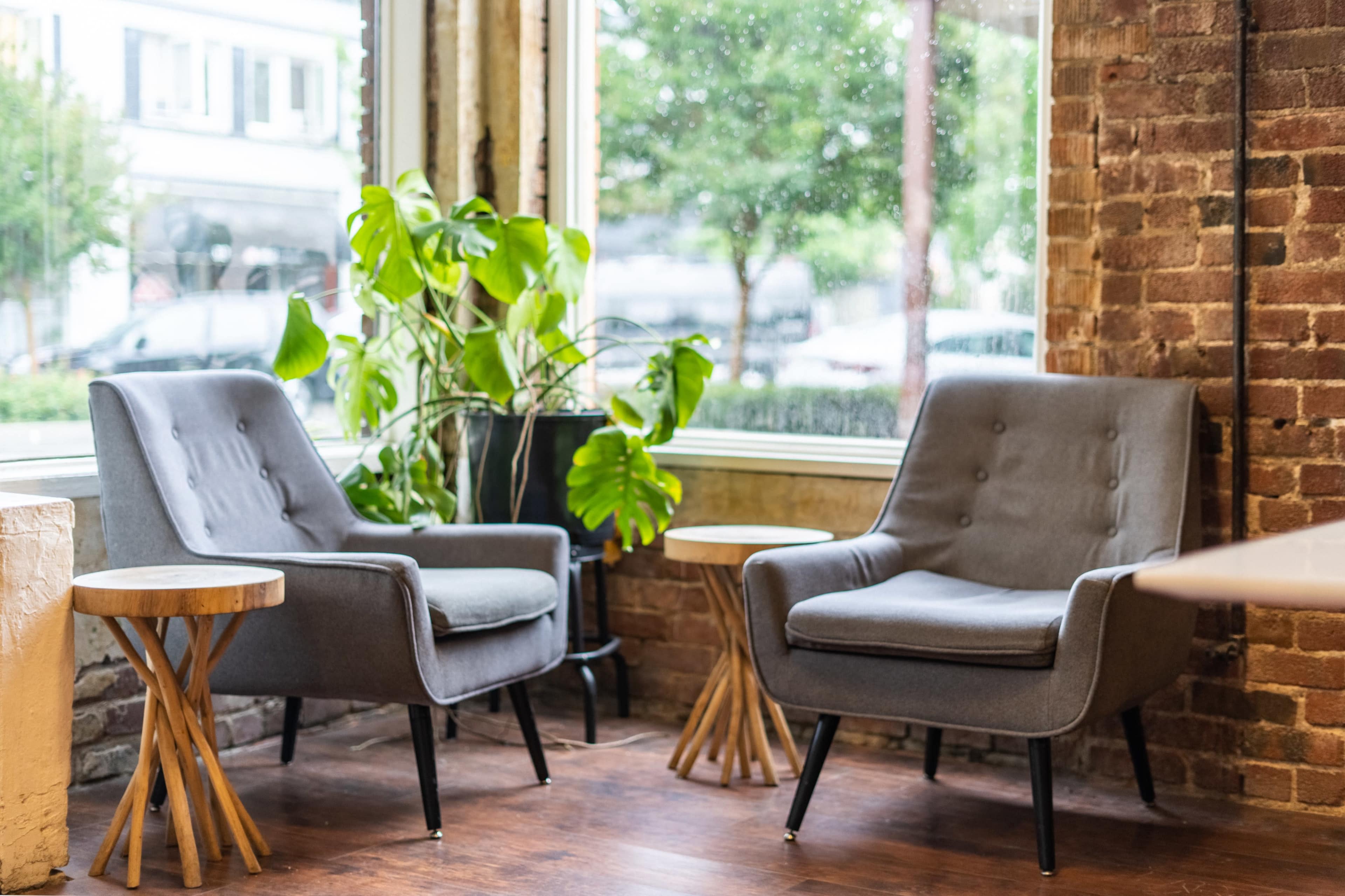 Two gray chairs with wooden legs are positioned beside small round tables in a cozy corner by a window with a view of greenery outside.