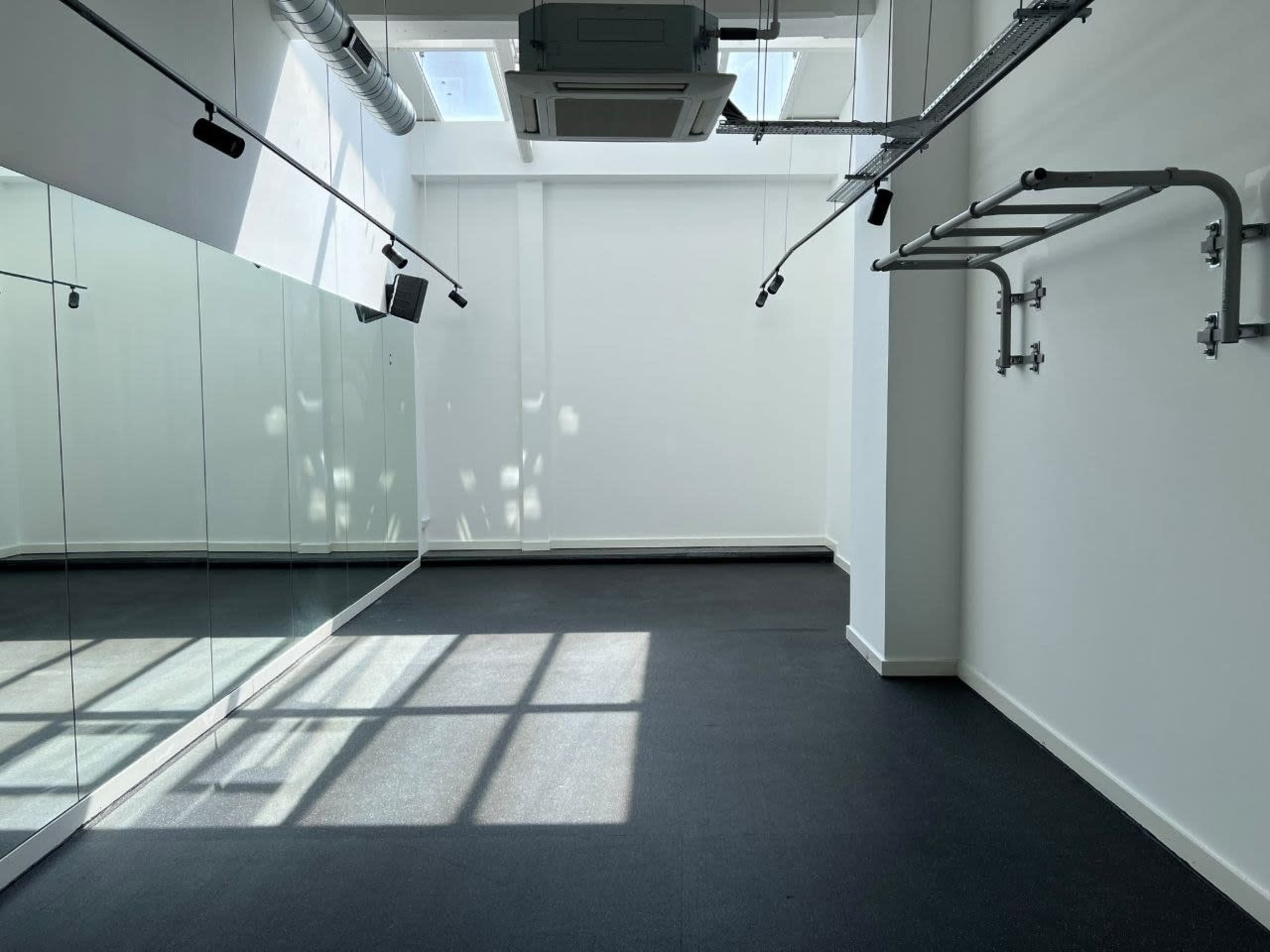 The image shows a spacious room with mirrored walls, a ceiling-mounted air conditioning unit, and a section of exposed piping, illuminated by natural light from overhead windows.