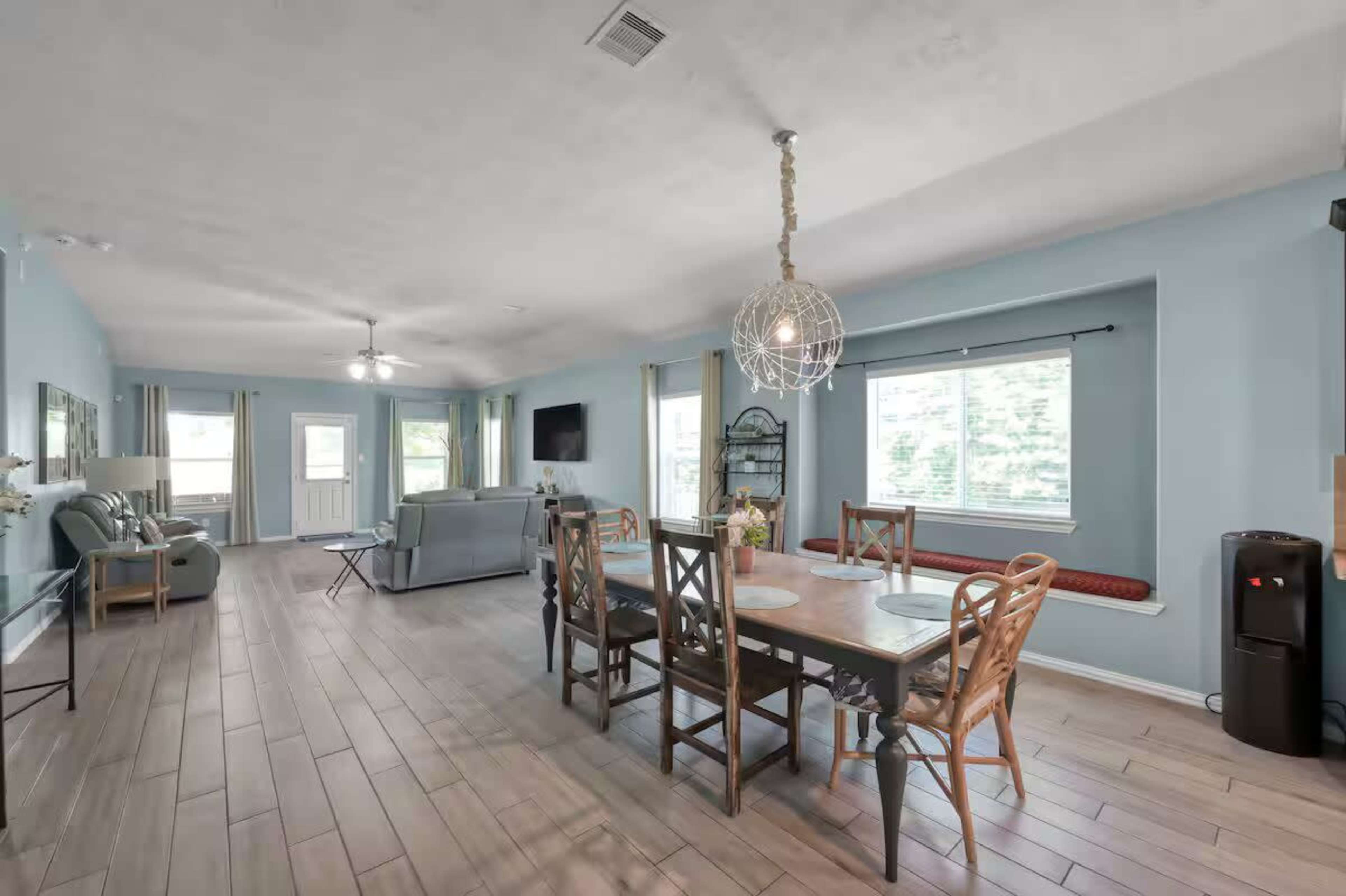 The image shows an open-concept living and dining area featuring a dining table with chairs, a couch, and large windows allowing natural light to brighten the space.