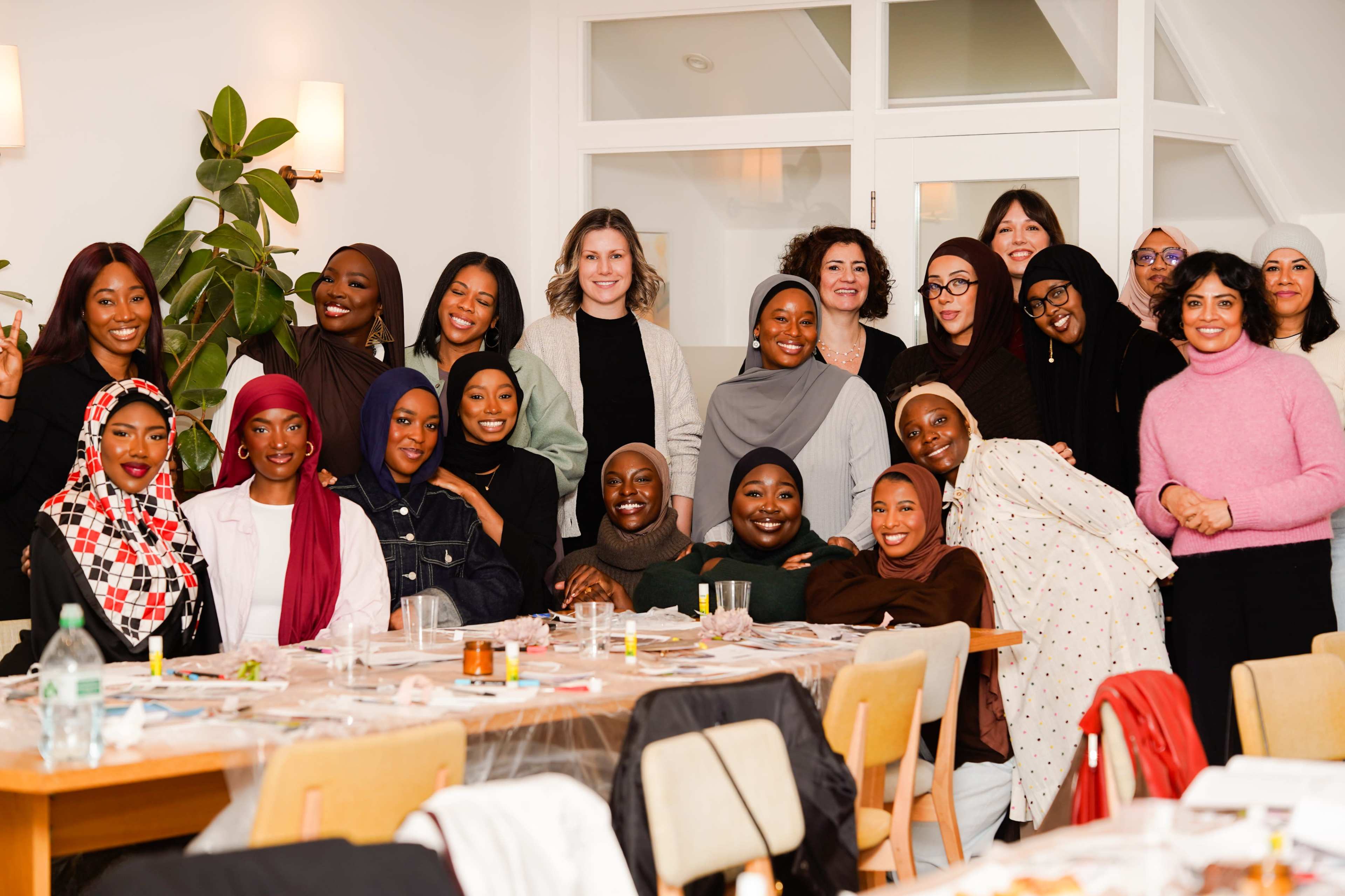 A diverse group of women is gathered together in a bright room, smiling for a photo in front of a table set with food and drinks.