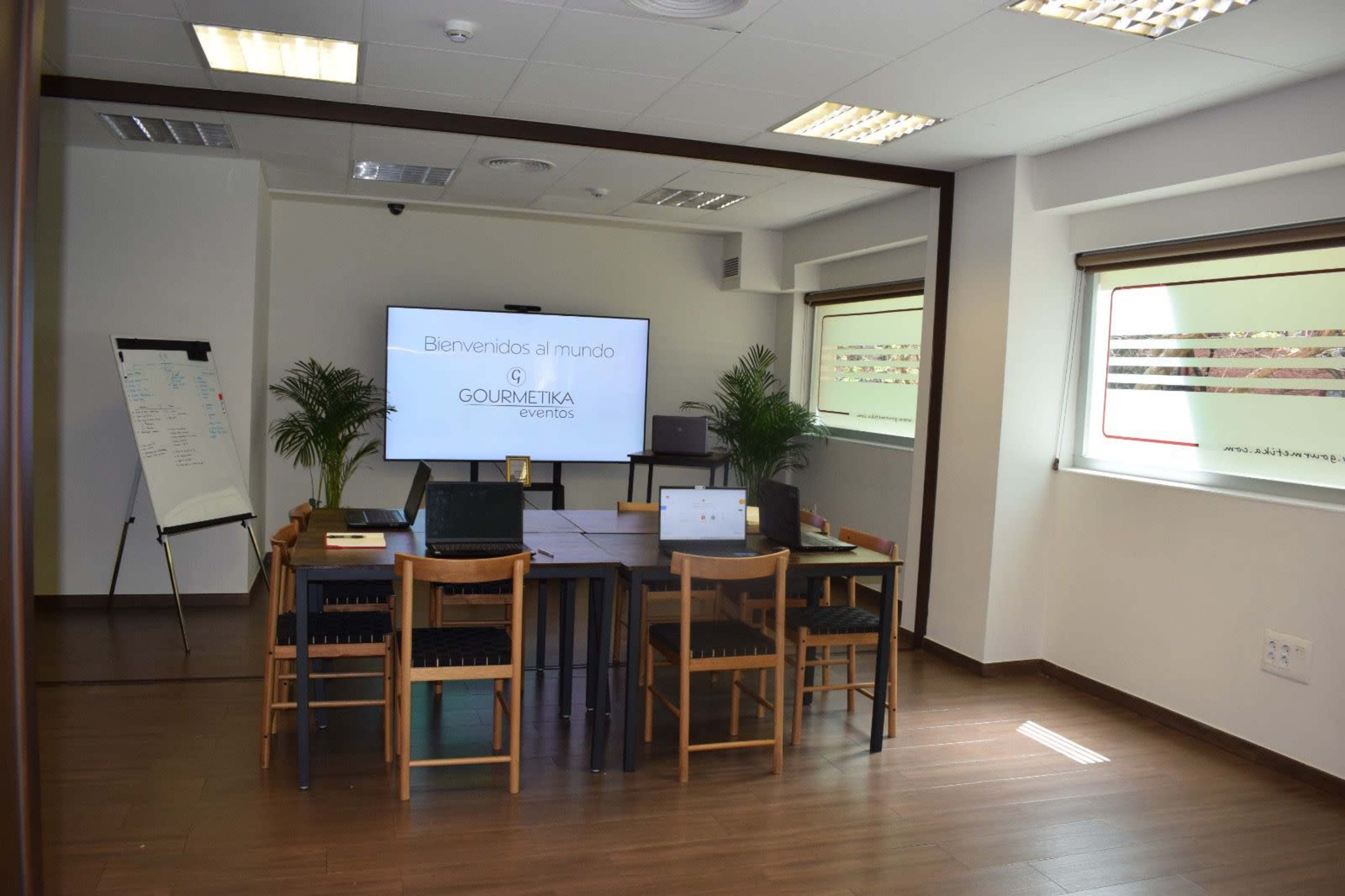 The image shows a modern conference room with a large screen displaying a welcome message, surrounded by a table and several chairs, along with laptops and a whiteboard.