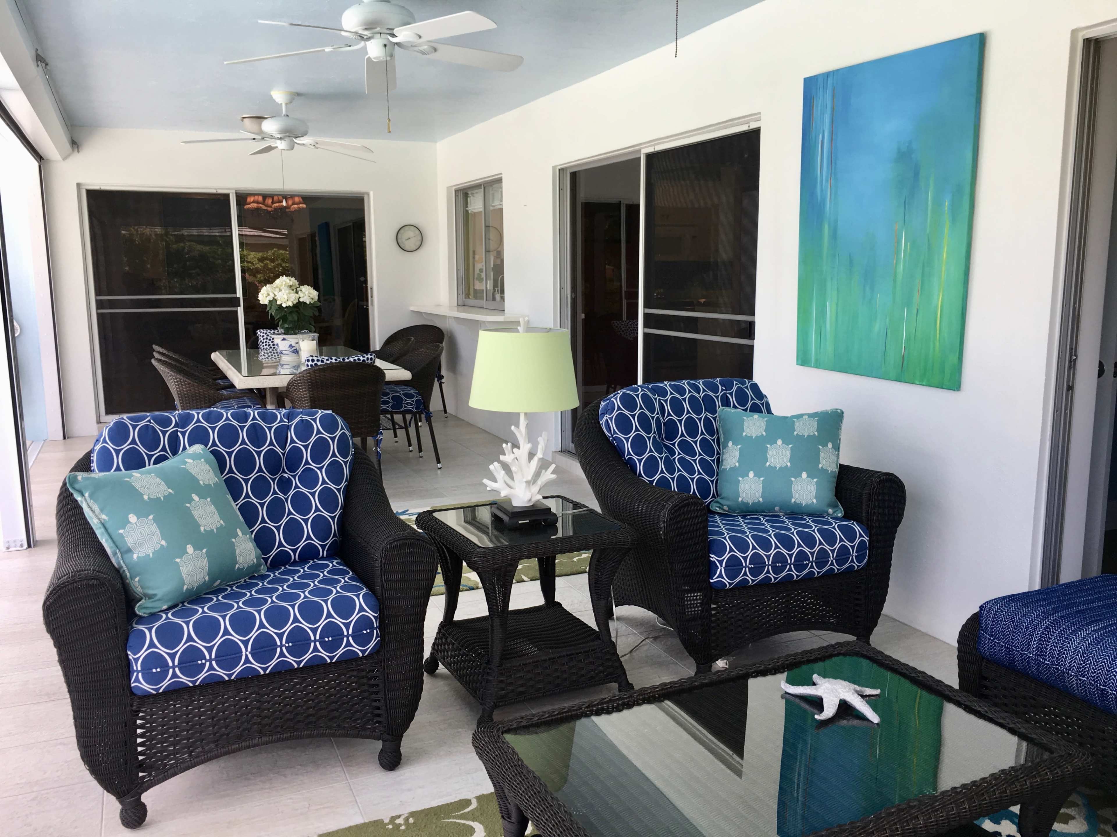A porch features two patterned chairs with a small table between them, a dining area in the background, and a large blue painting on the wall.