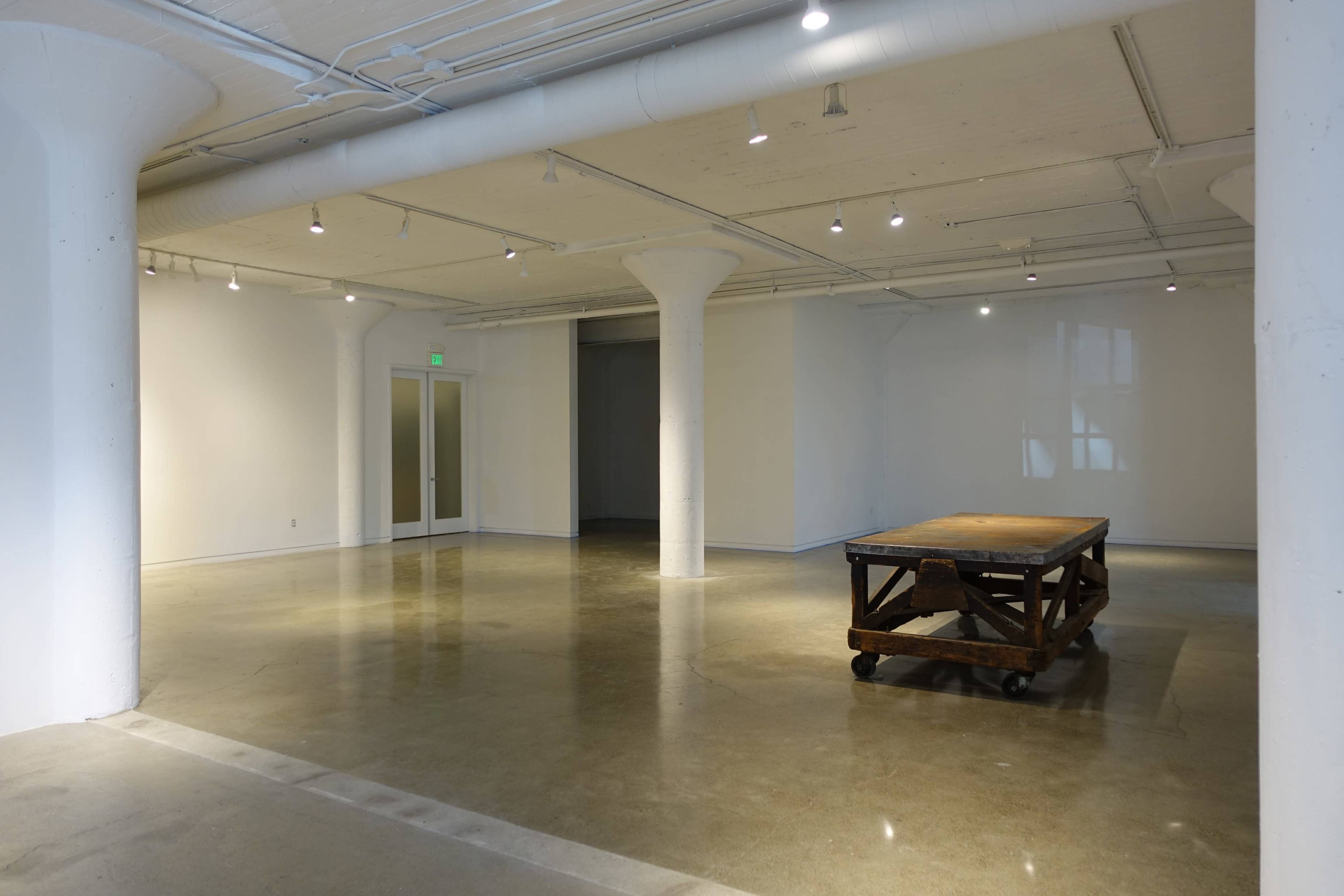 The image shows a spacious, minimalist room with polished concrete floors, white walls, and a wooden table on wheels in the center.