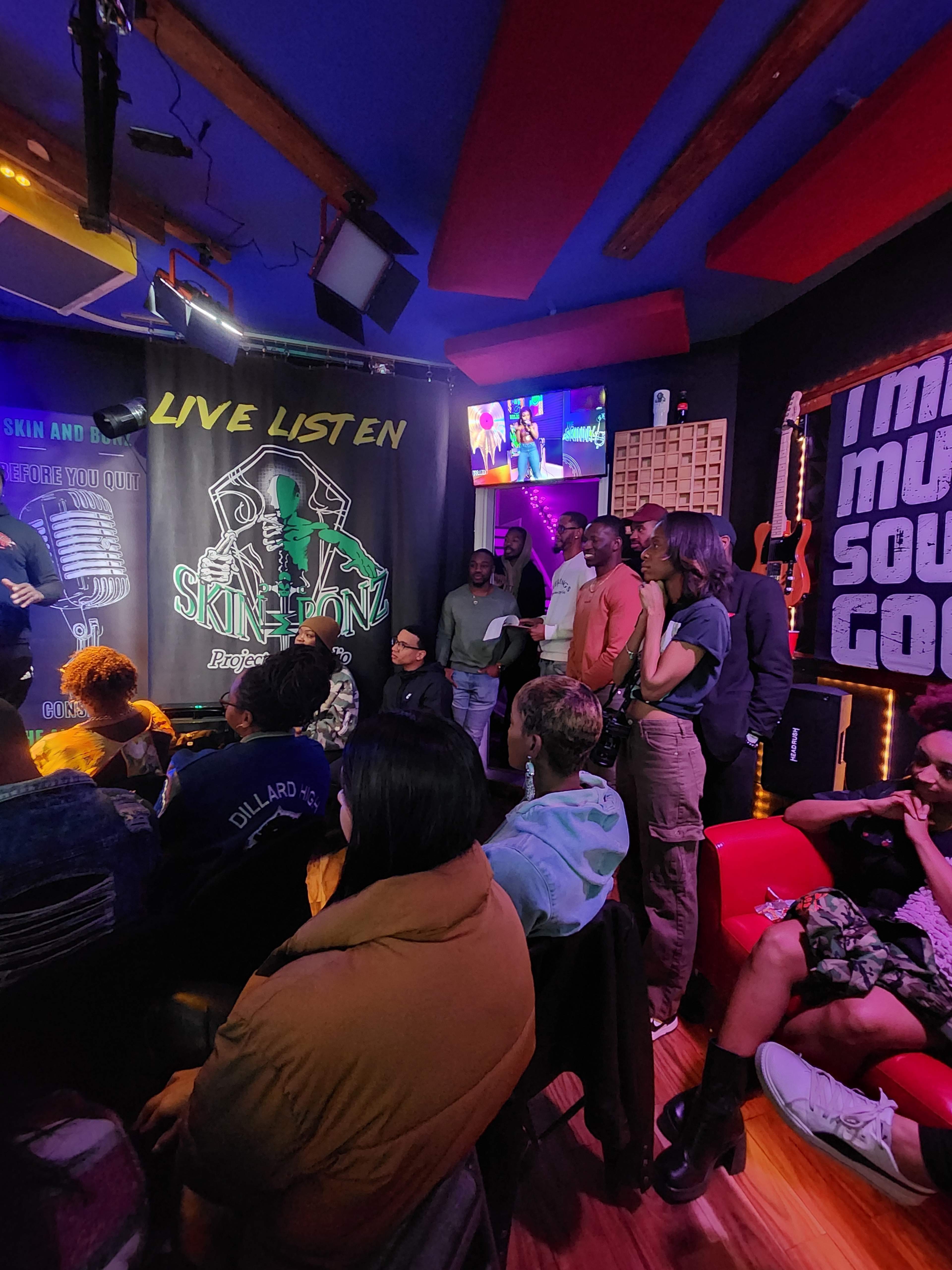 A group of people stands in a dimly lit venue with musical decorations, while a performer is on stage.