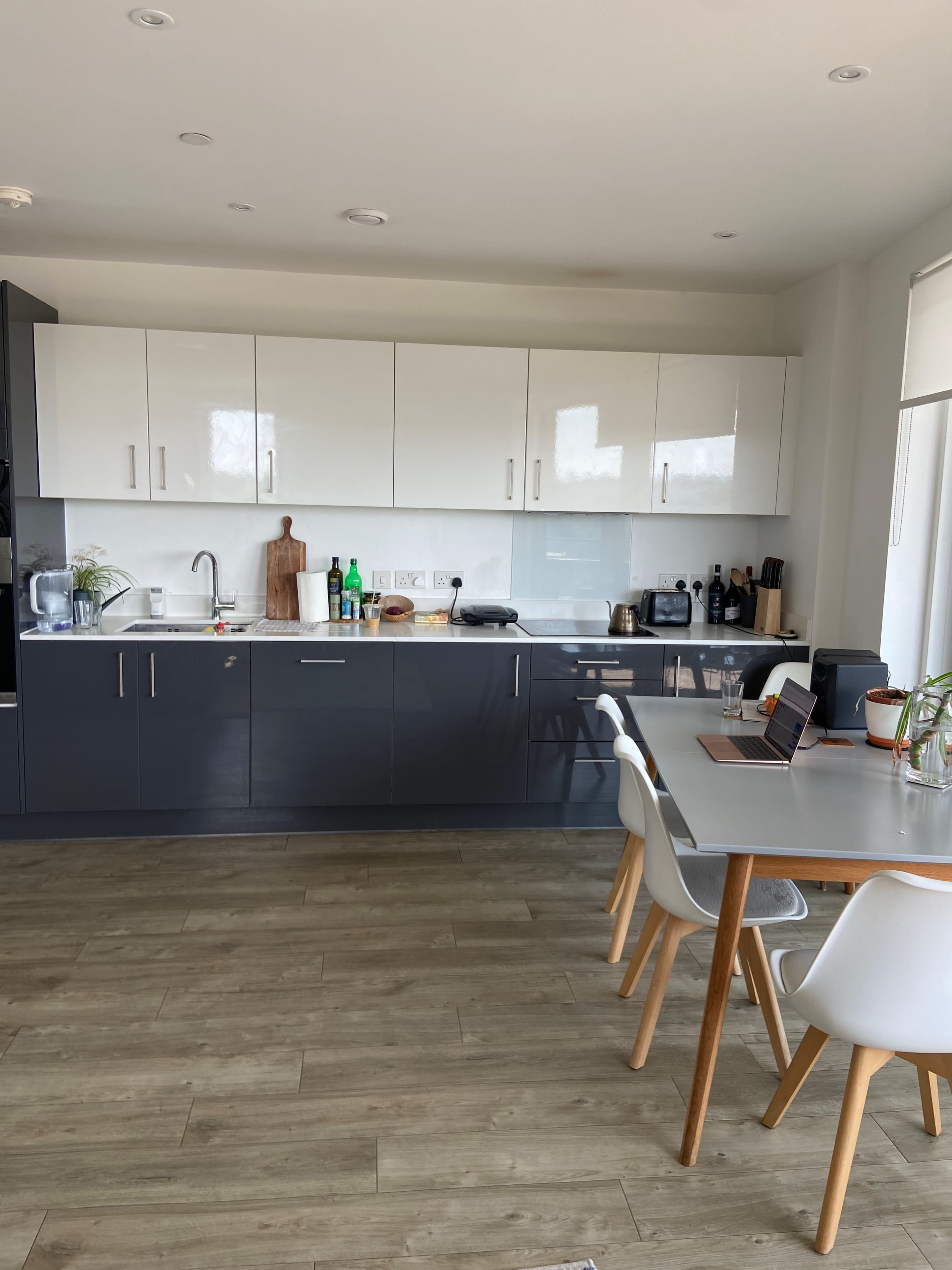 The image shows a modern kitchen with white and dark cabinetry, a wooden dining table with white chairs, and various kitchen appliances on the counter.