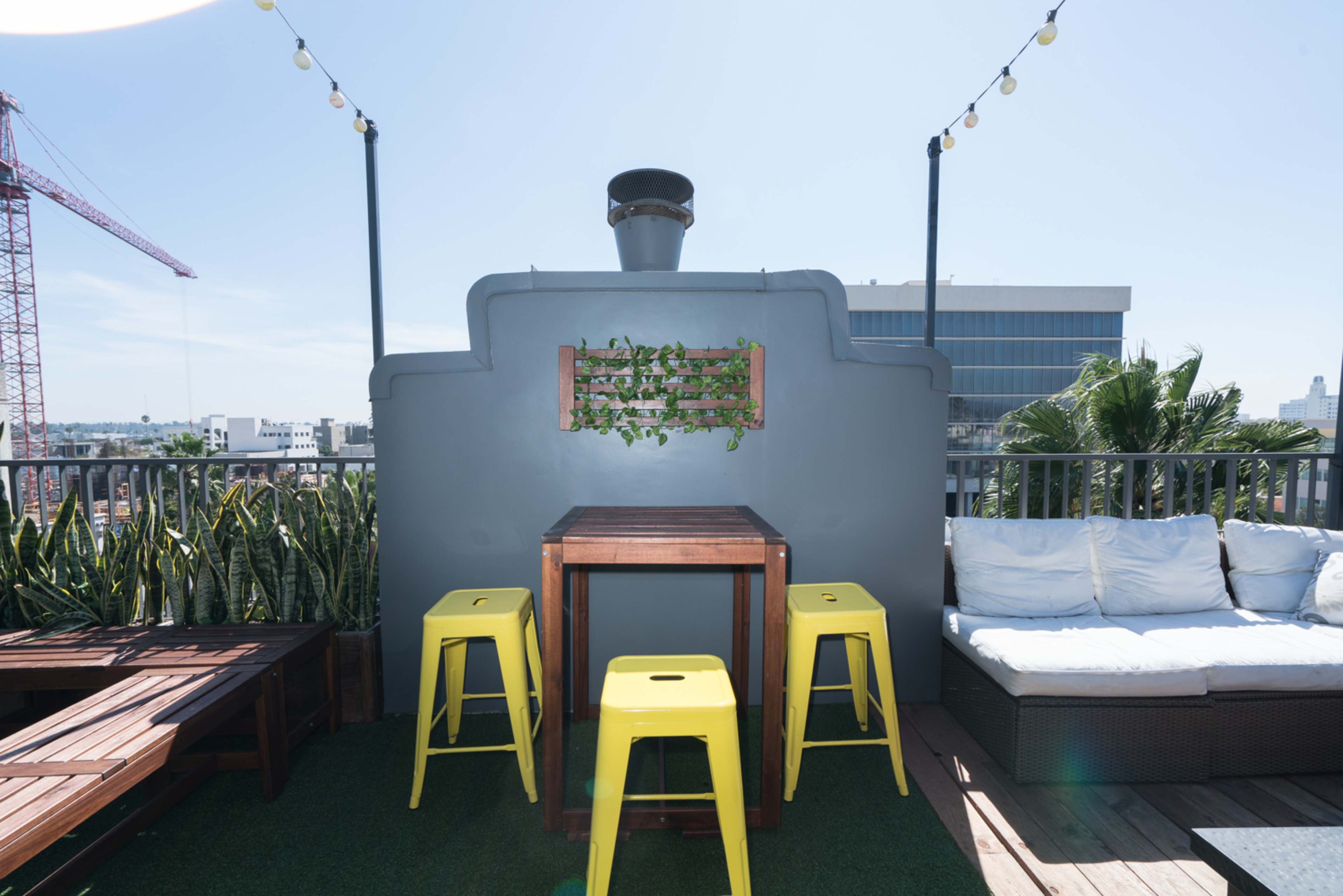 A rooftop area features a wooden table with yellow stools, a gray wall with a plant display, and a backdrop of city buildings under a clear sky.