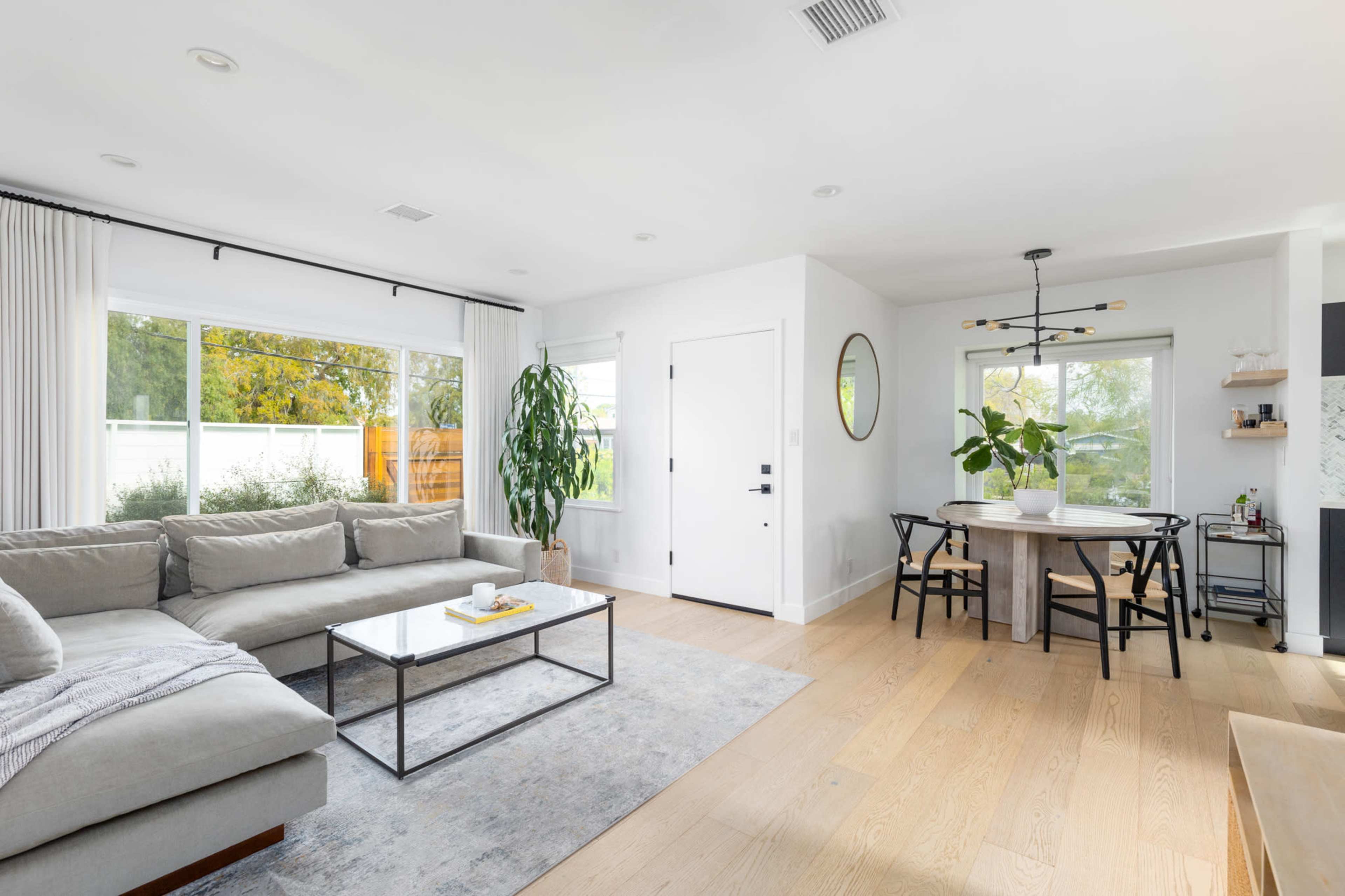 A modern living room and dining area with a sectional sofa, a coffee table, a round dining table, and large windows that offer natural light.
