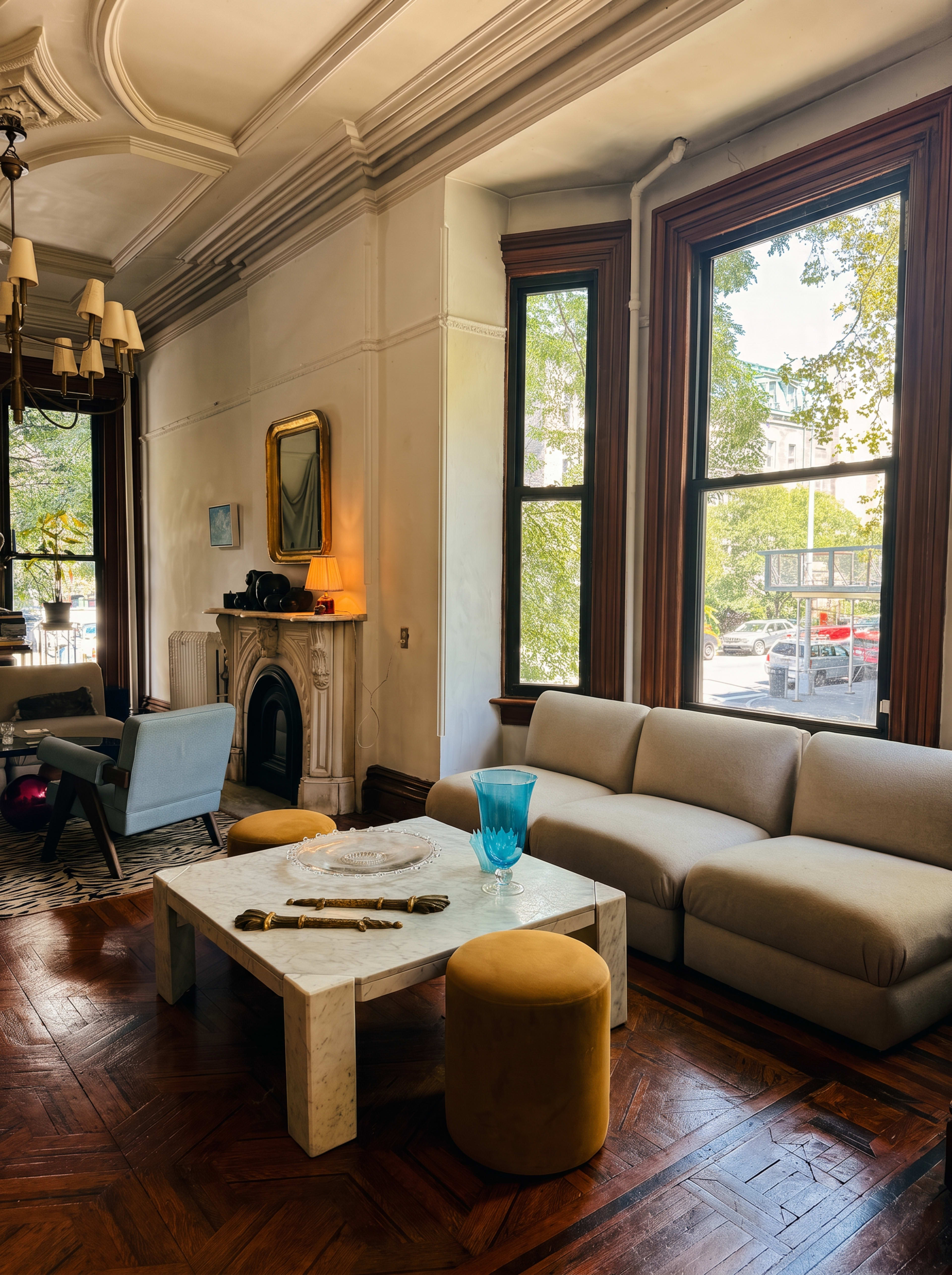 The image shows a well-lit living room featuring a beige sofa, a marble coffee table, and large windows with wooden trim.