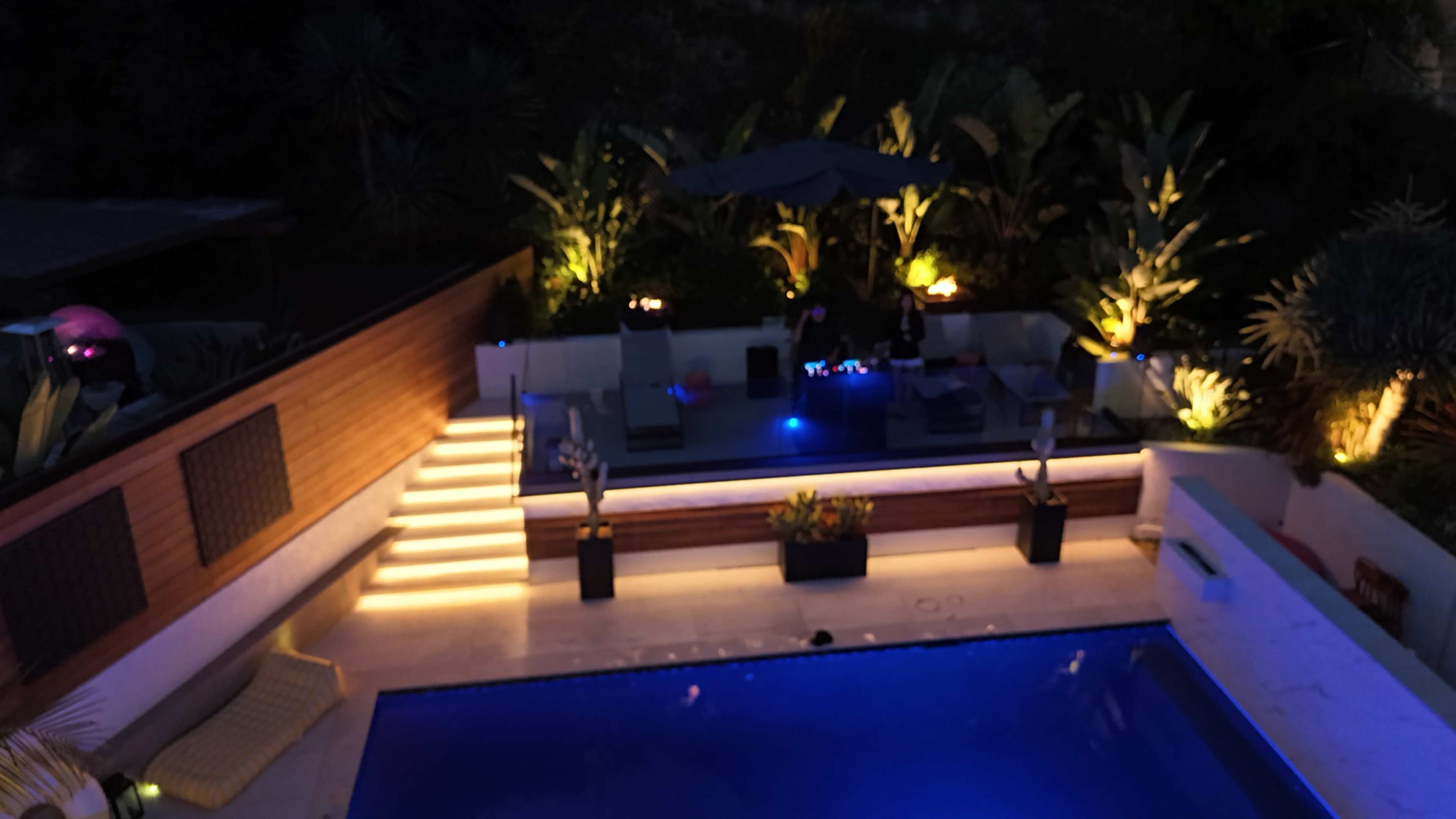 The image shows a nighttime outdoor pool area with a lounge seating arrangement, illuminated by various lights and surrounded by tropical plants.