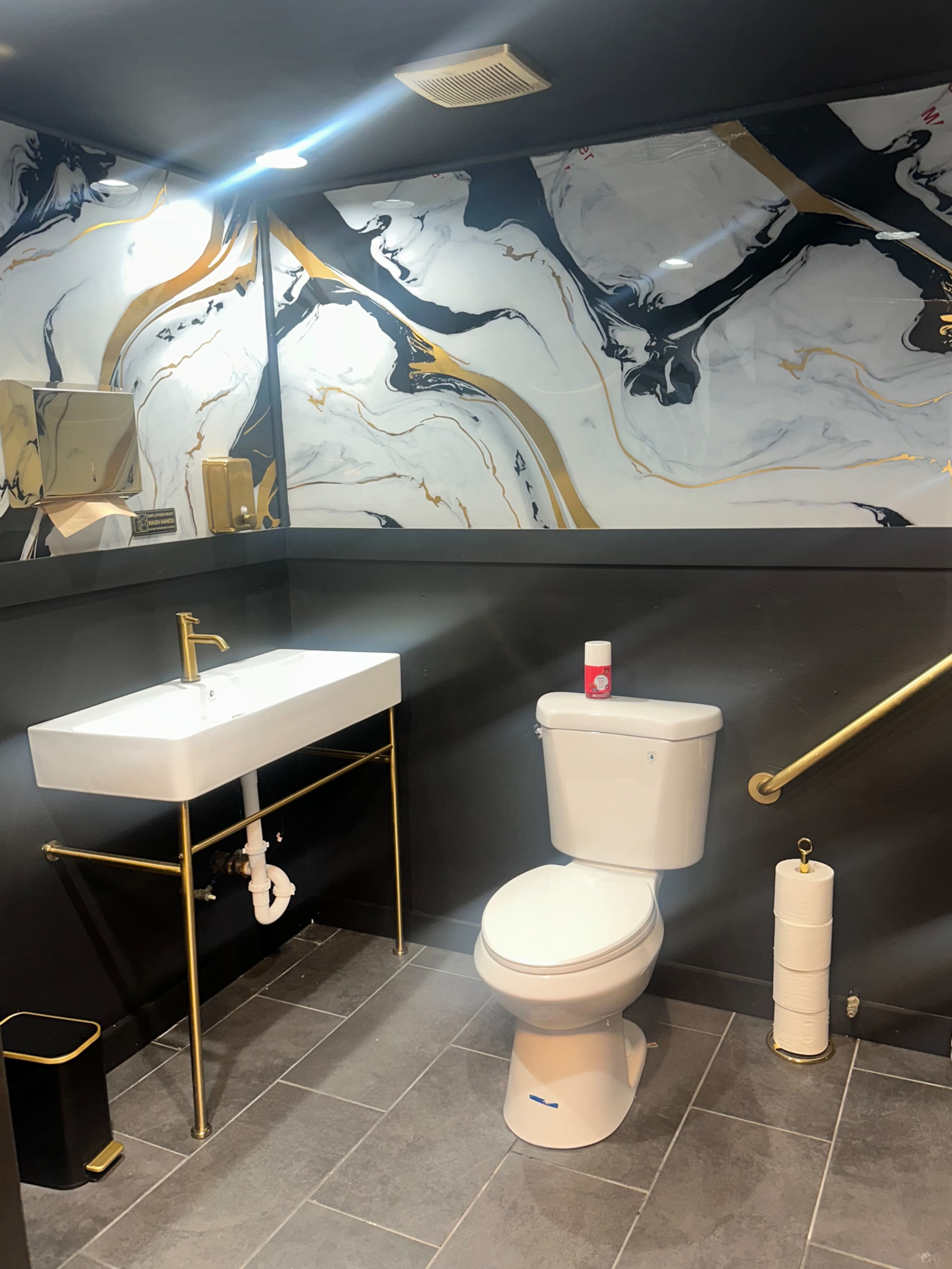 The image shows a modern bathroom featuring a white sink with gold accents, a white toilet, dark gray walls with a marbled design, and gray tiled flooring.
