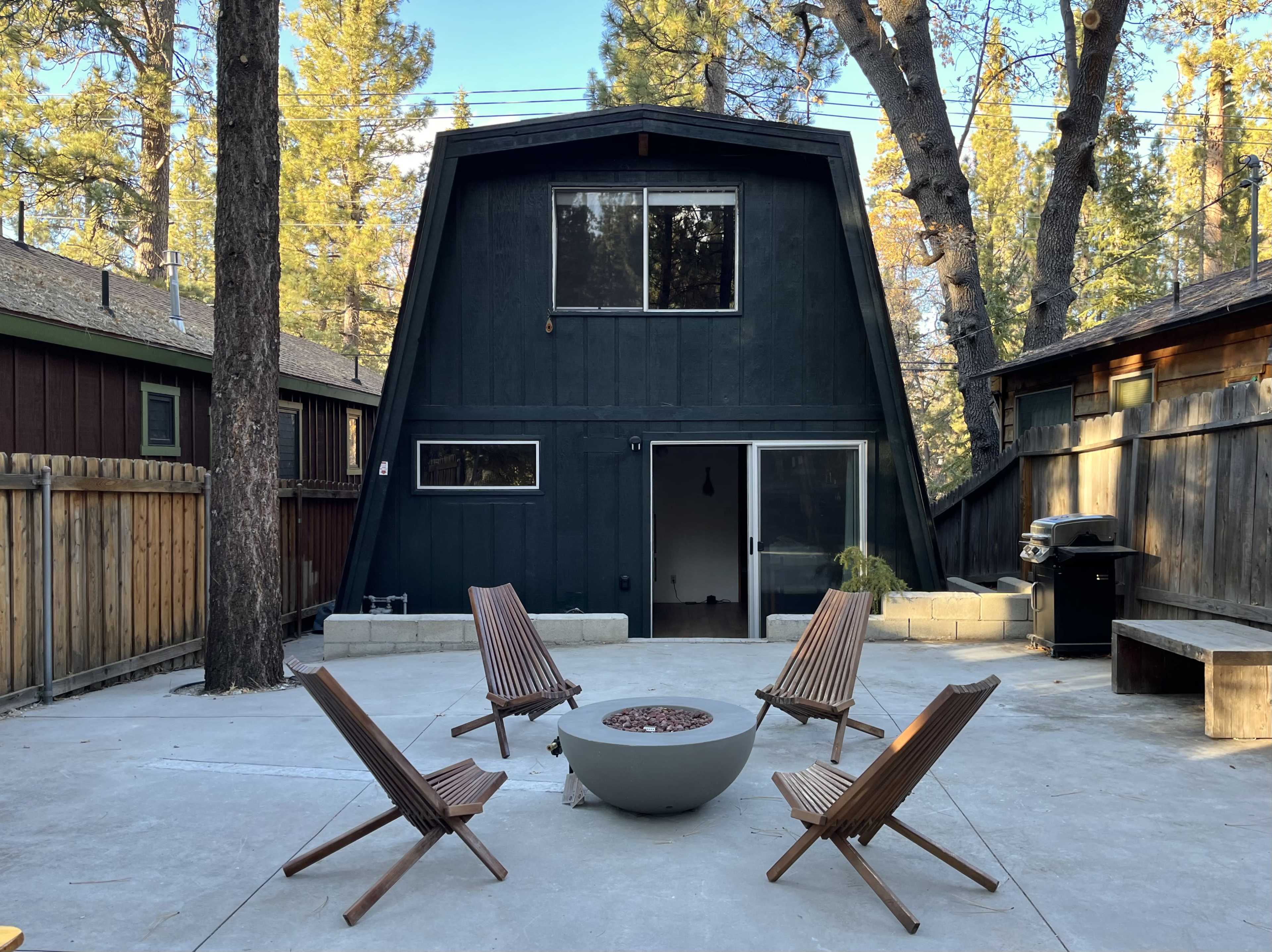 A modern A-frame house sits in a forested area, surrounded by gravel and wooden chairs arranged around a fire pit.