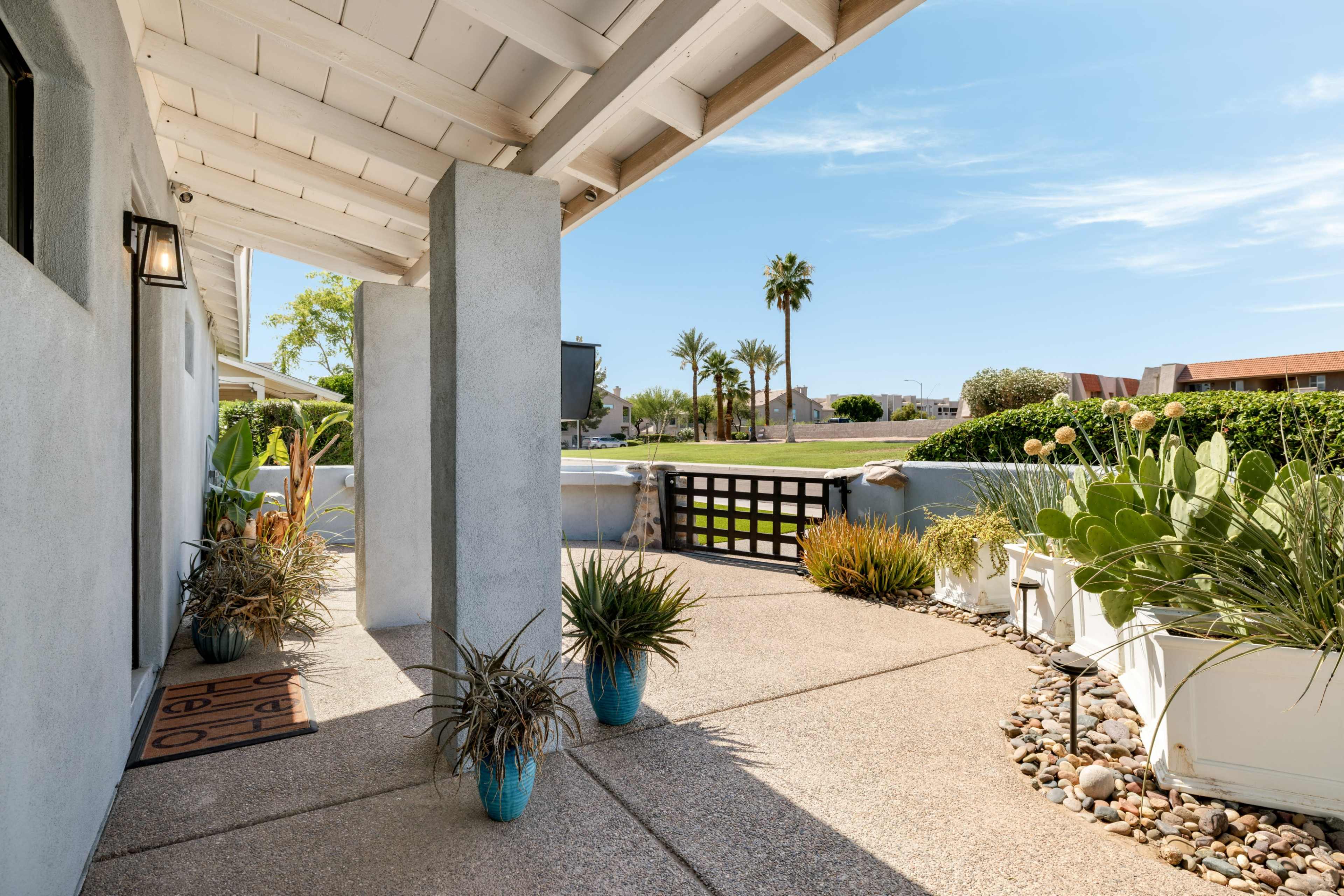 The image shows a covered porch with potted plants and a view of a landscaped yard featuring palm trees and a white fence.