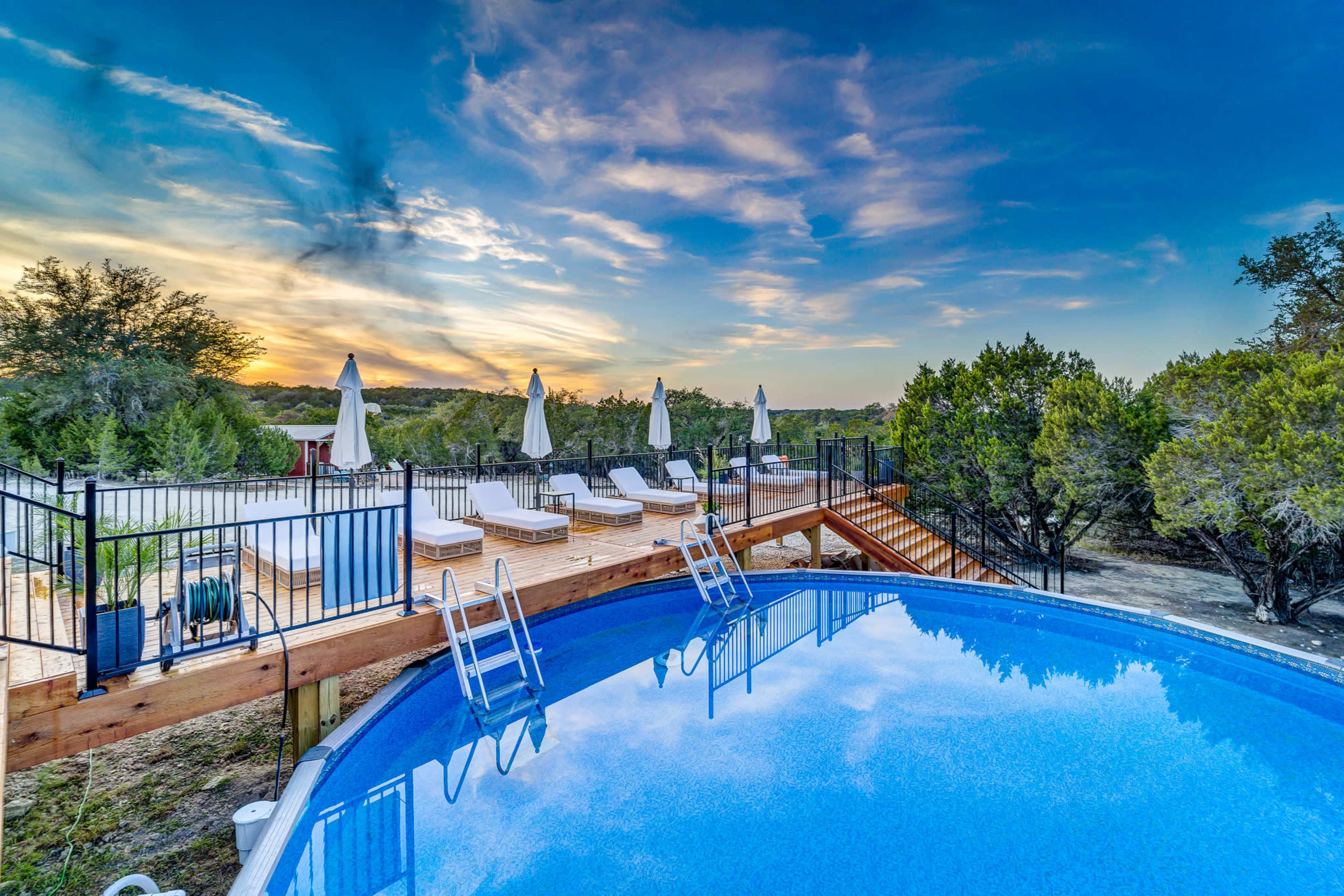 The image shows a swimming pool surrounded by wooden decking and lounge chairs, with umbrellas in the background against a sunset sky.