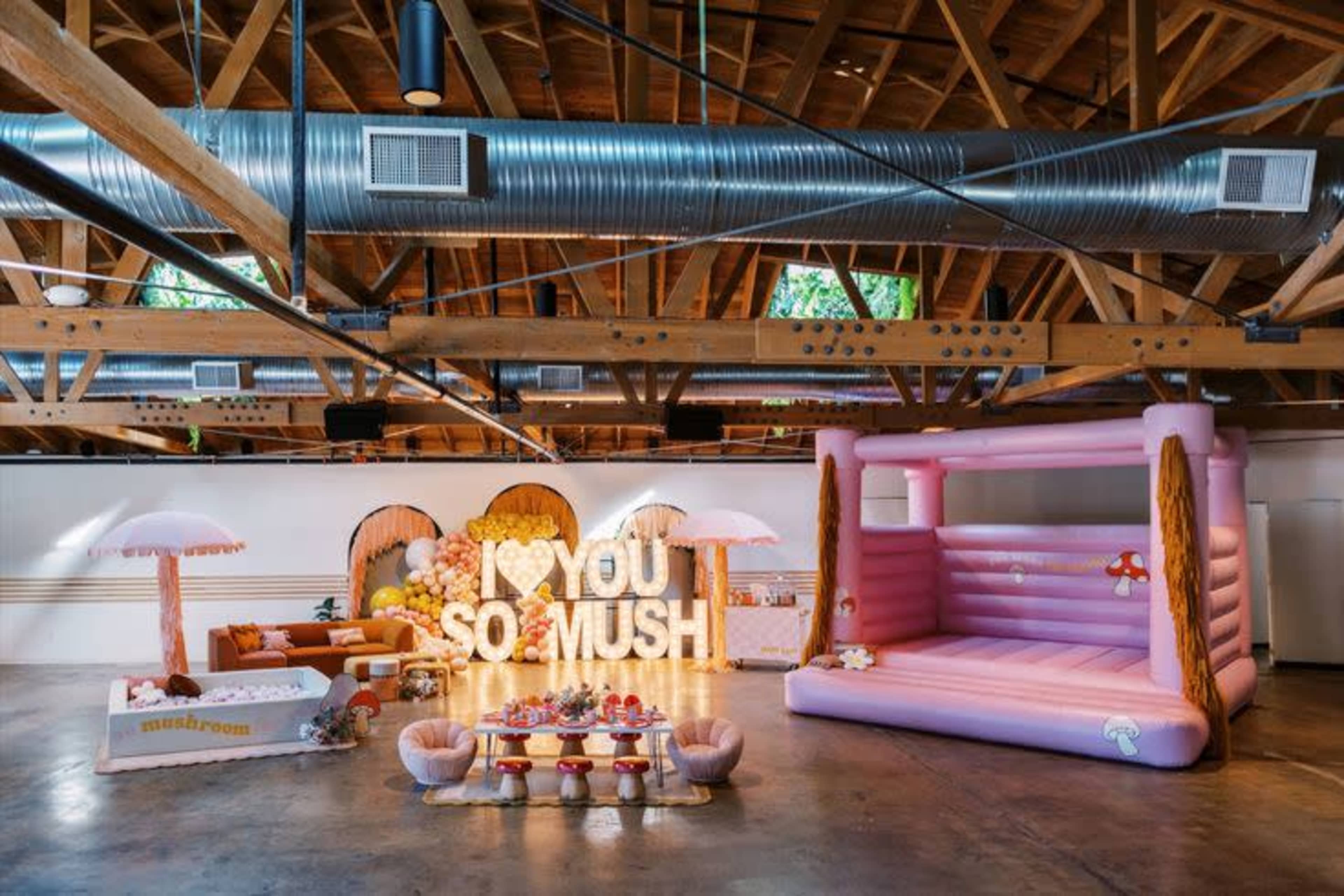 The image shows a spacious indoor venue decorated with pastel-colored furniture, a pink inflatable bounce house, and illuminated letters that read "I KYOU SO MUCH."