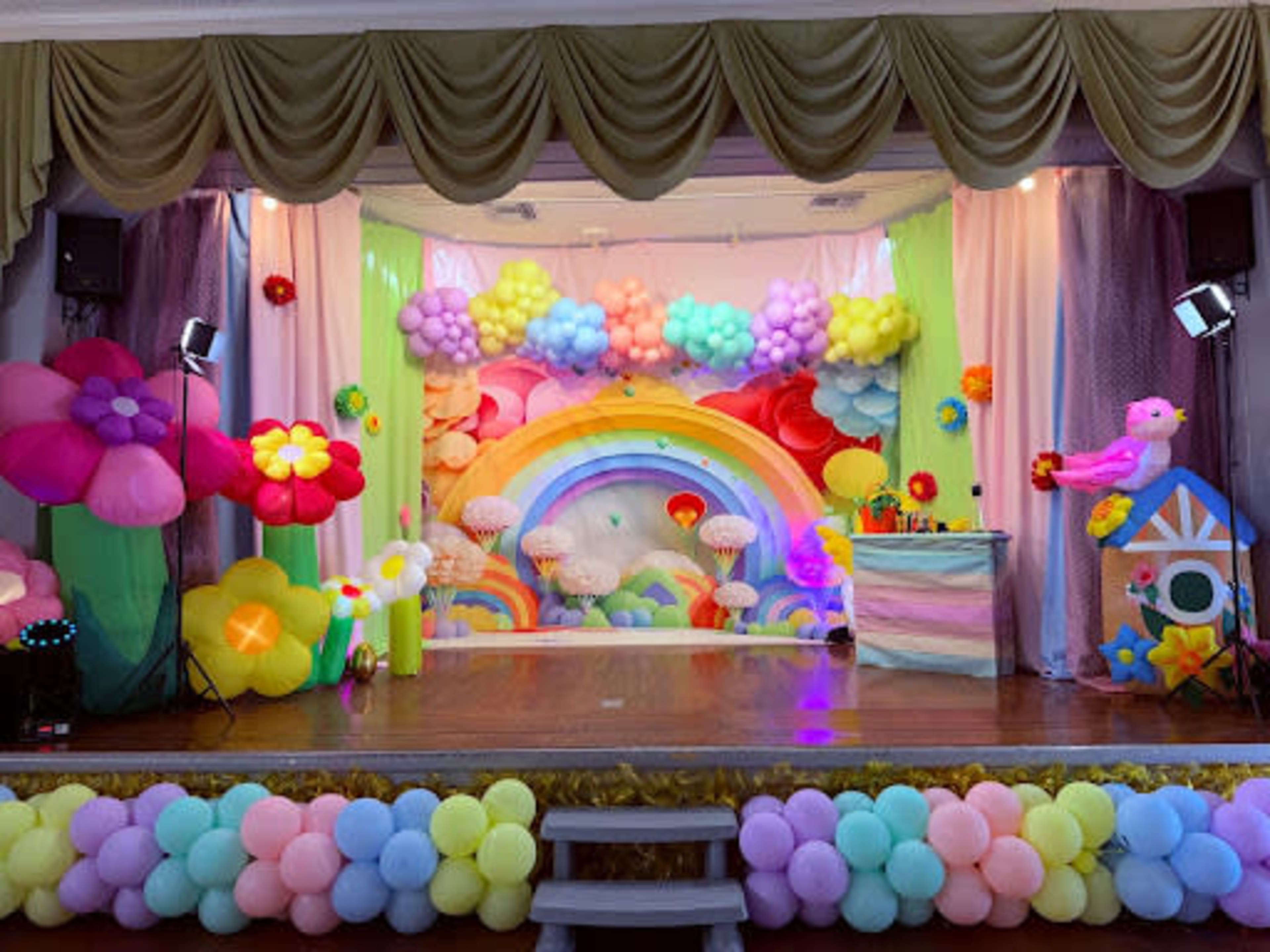The image shows a colorful stage decorated with balloons in various shapes and colors, featuring a rainbow backdrop, floral elements, and a small decorated table.