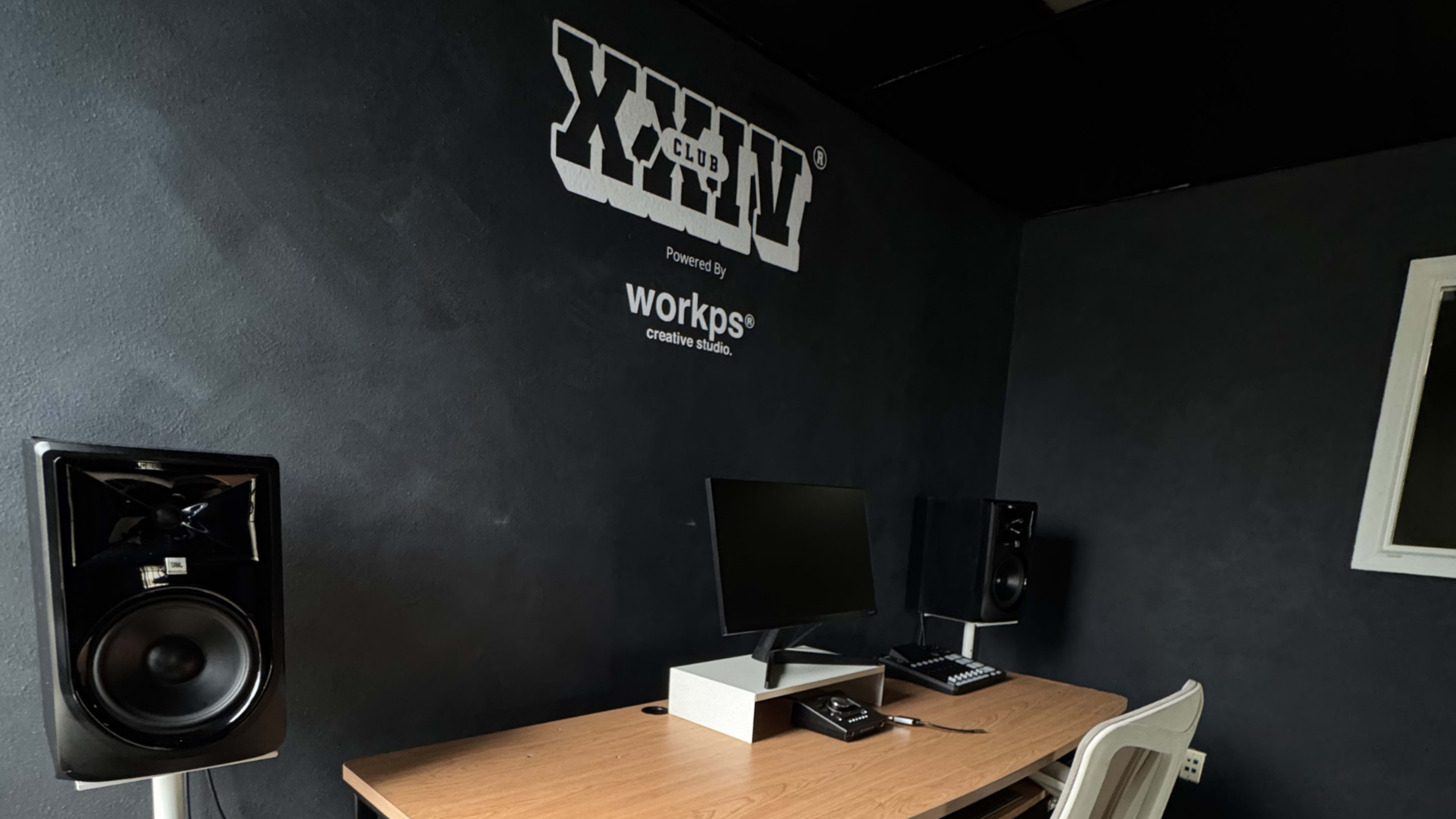 The image shows a minimalist workspace featuring a wooden desk with a computer and audio monitors against a dark wall with the text "XXIV Club" and "Powered by workps creative studio."