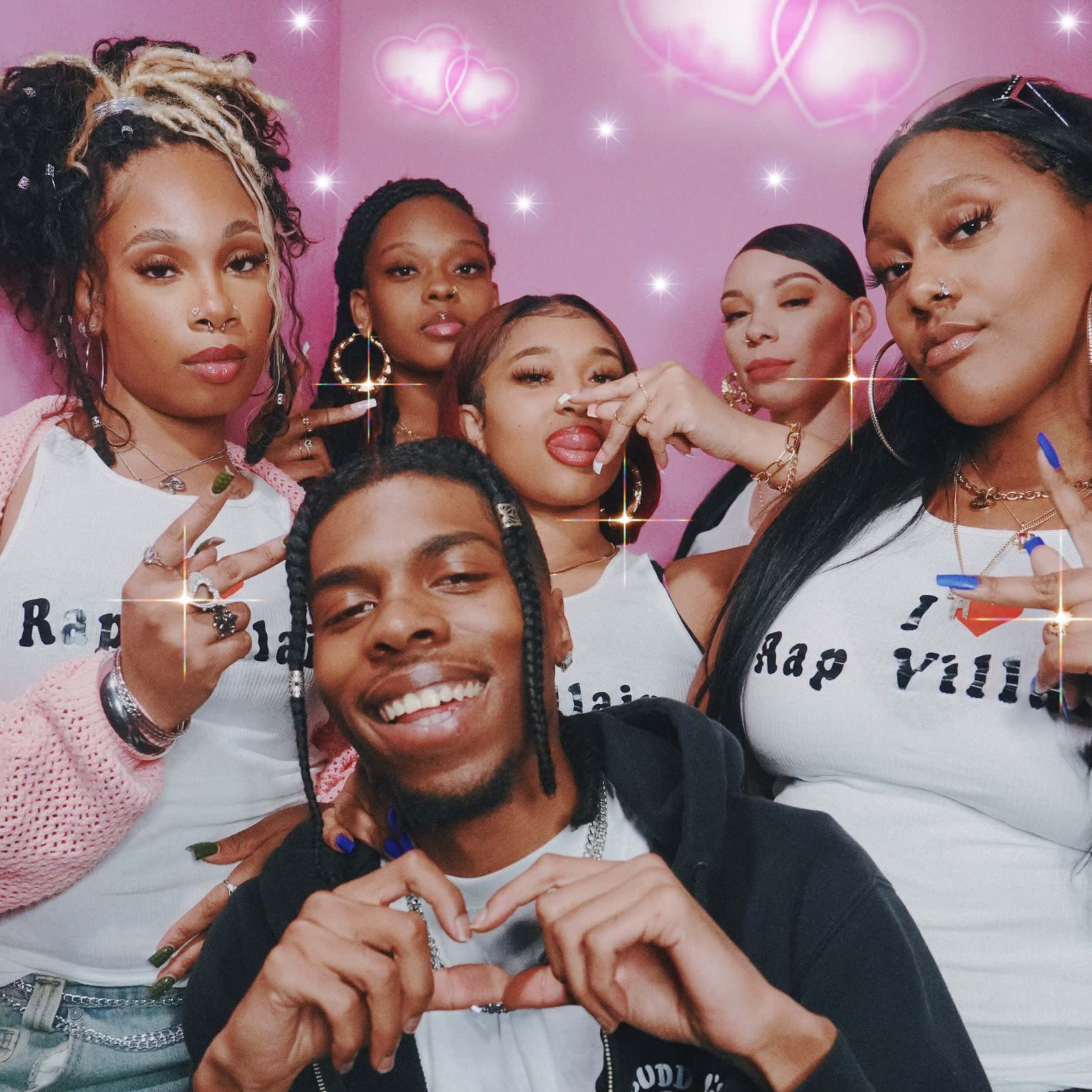 A group of six individuals poses together against a pink backdrop with decorative hearts, wearing matching shirts that say "Rap Villa."