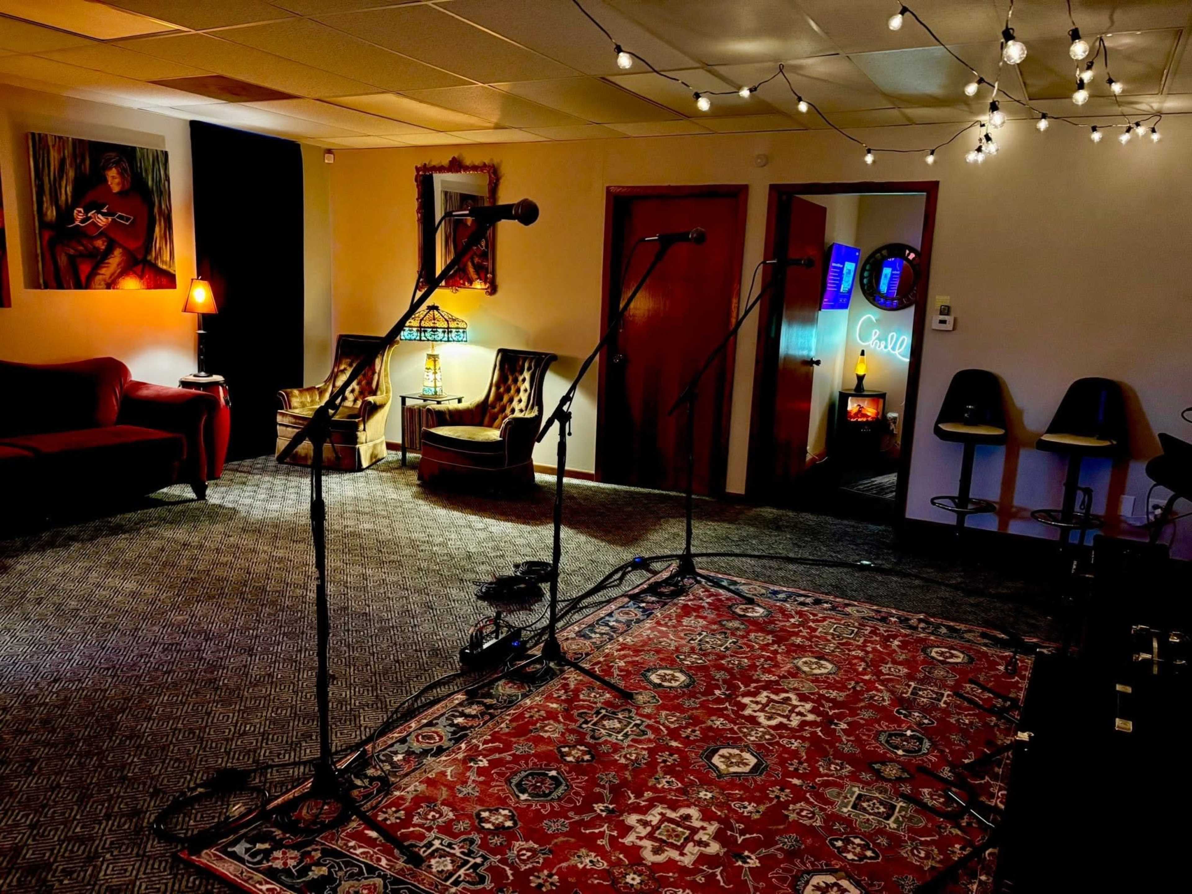 The room features a cozy setup with microphones on stands, patterned rugs, and vintage furniture illuminated by soft lighting and string lights.