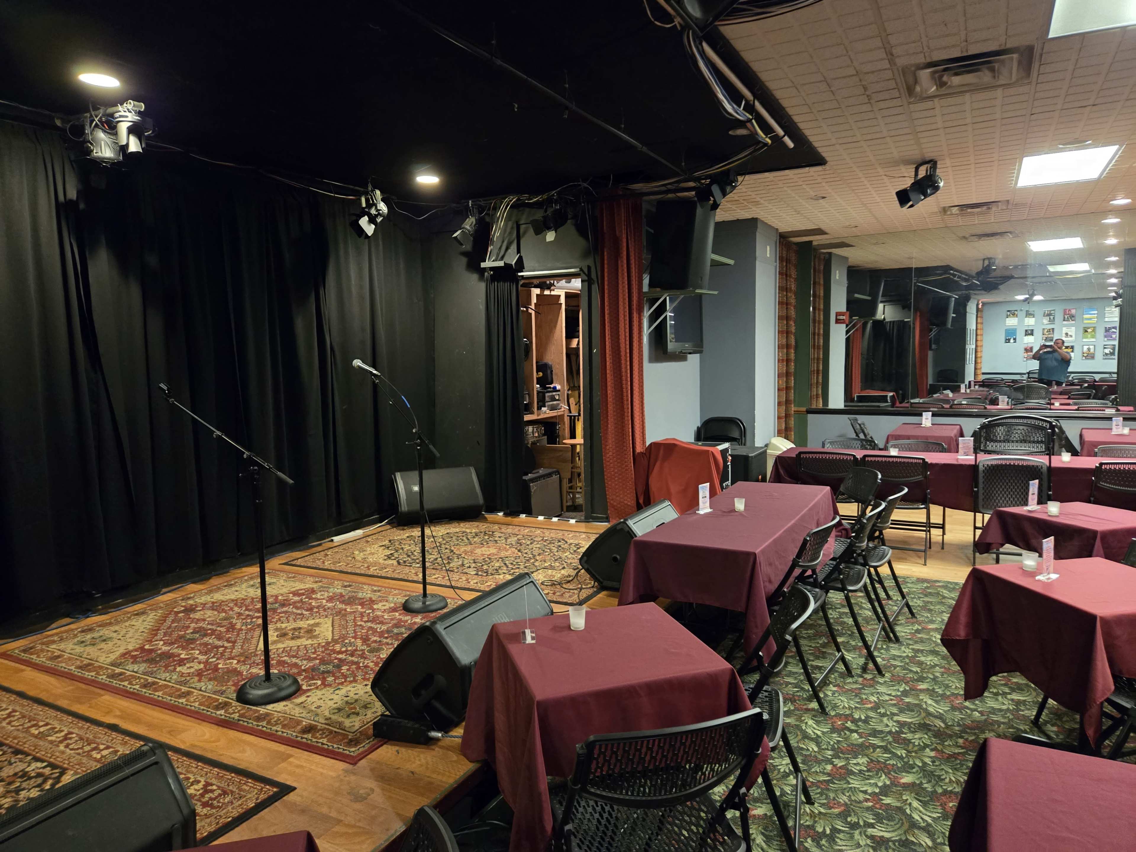 The image shows a stage with two microphones and amplifiers beside an array of tables covered with maroon tablecloths in a dimly lit performance space.