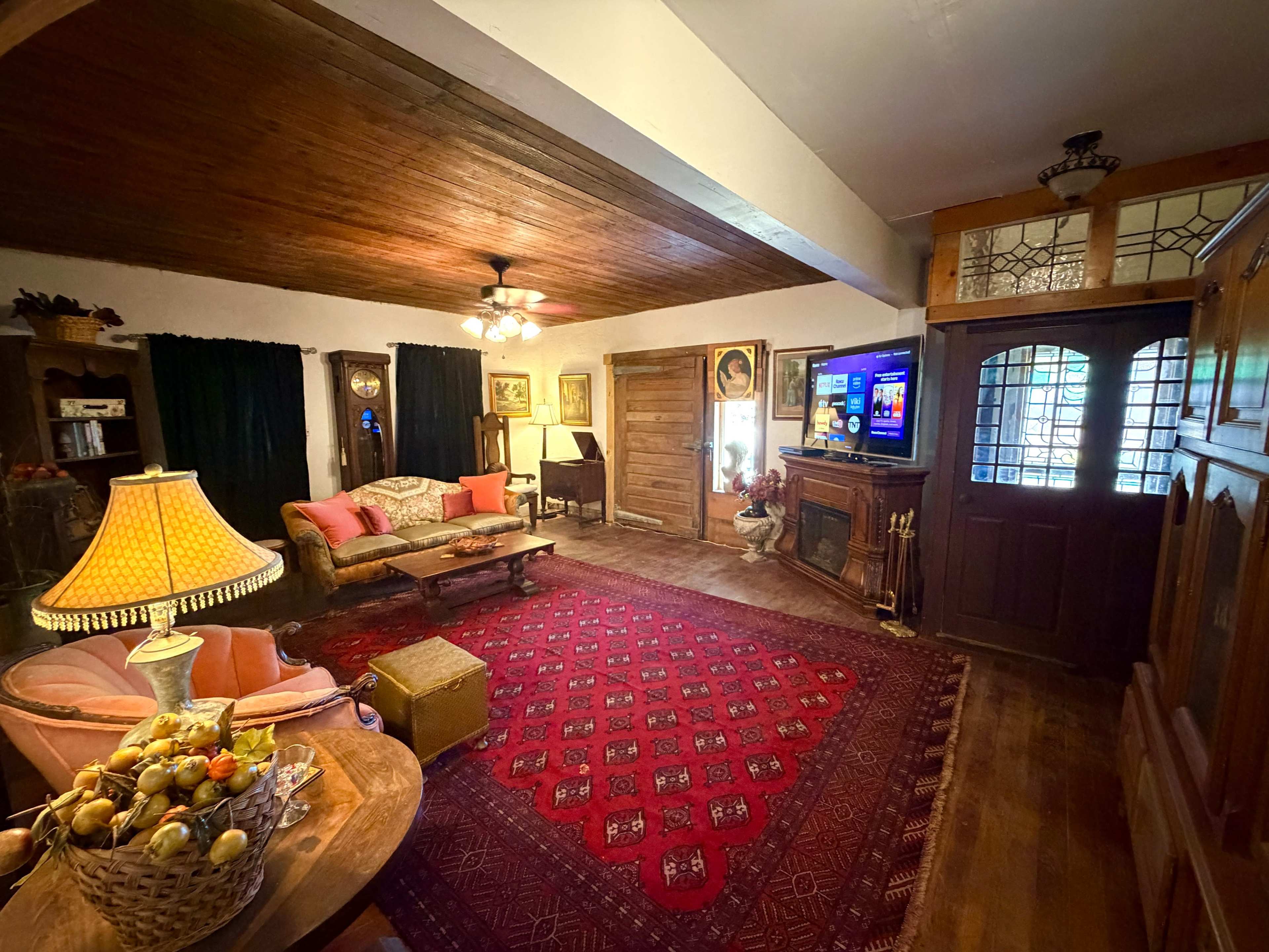 The interior of a cozy living room features a vintage sofa, a large red area rug, a television mounted on the wall, and wooden furnishings with decorative items.