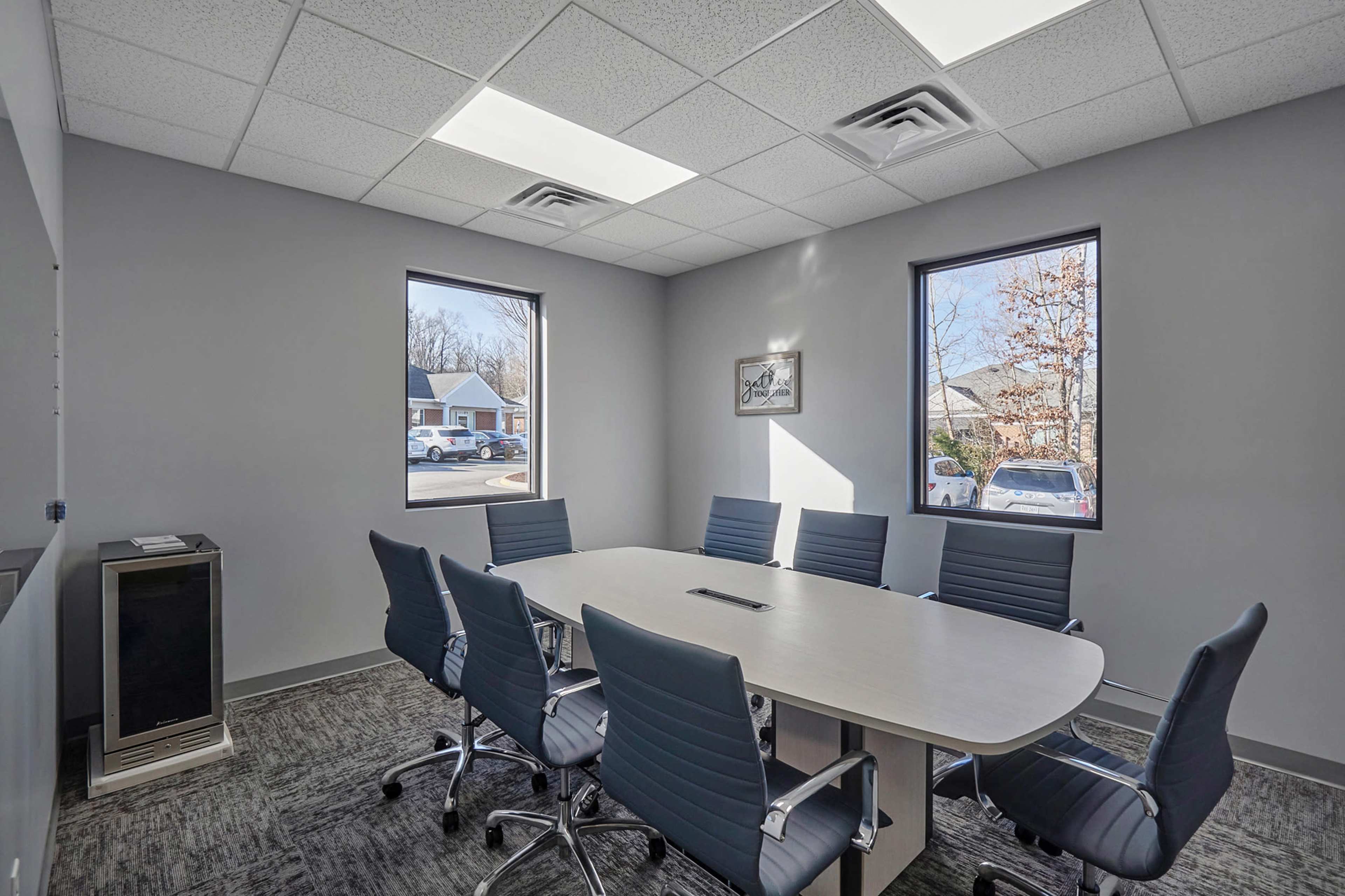 A conference room features a large oval table surrounded by gray rolling chairs, with windows providing views of an outside parking area.