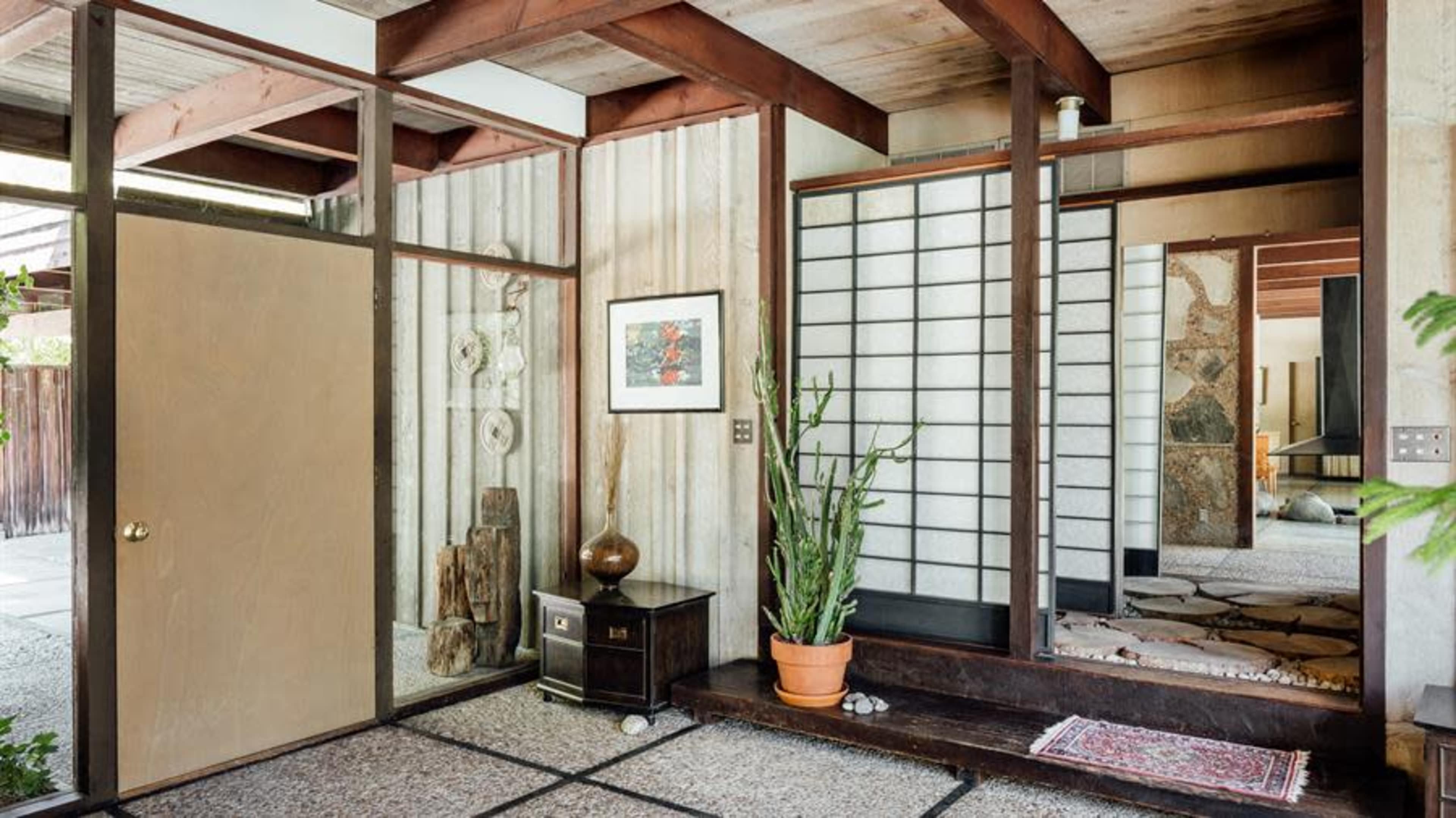 A modern interior entrance with a combination of glass walls, wooden beams, and a stone floor, featuring a door and decorative plants.