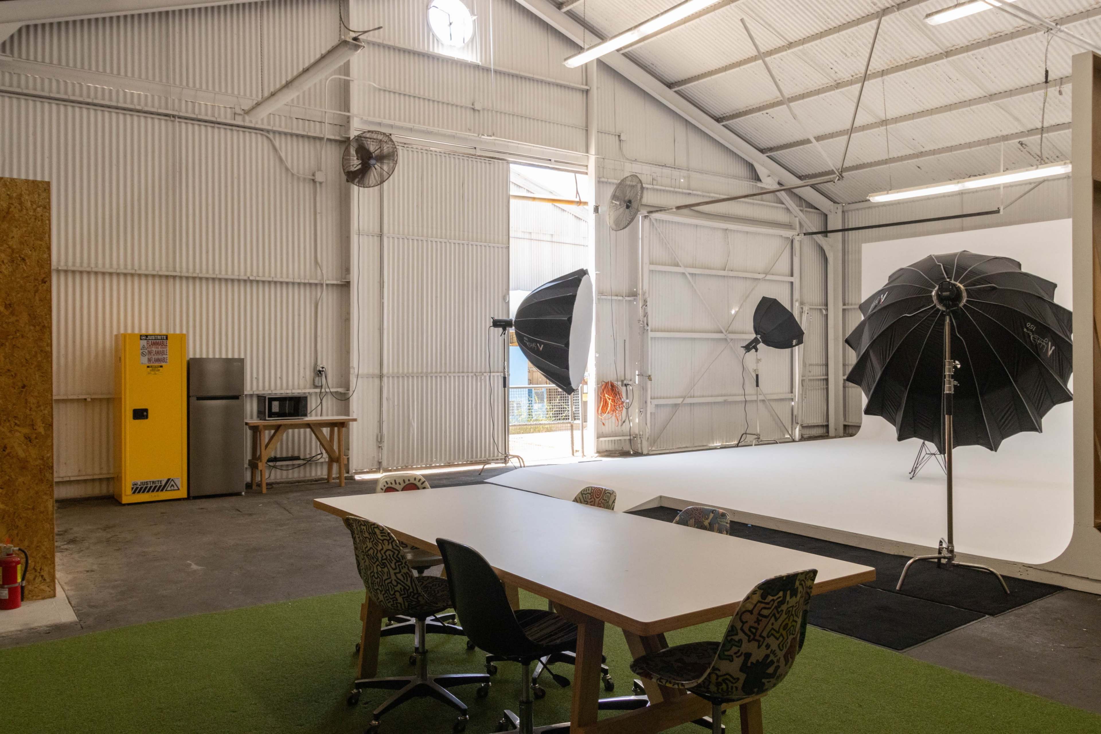 The scene features a bright industrial photography studio with a table and chairs, a yellow fridge, and large softbox lights set up on a white backdrop.