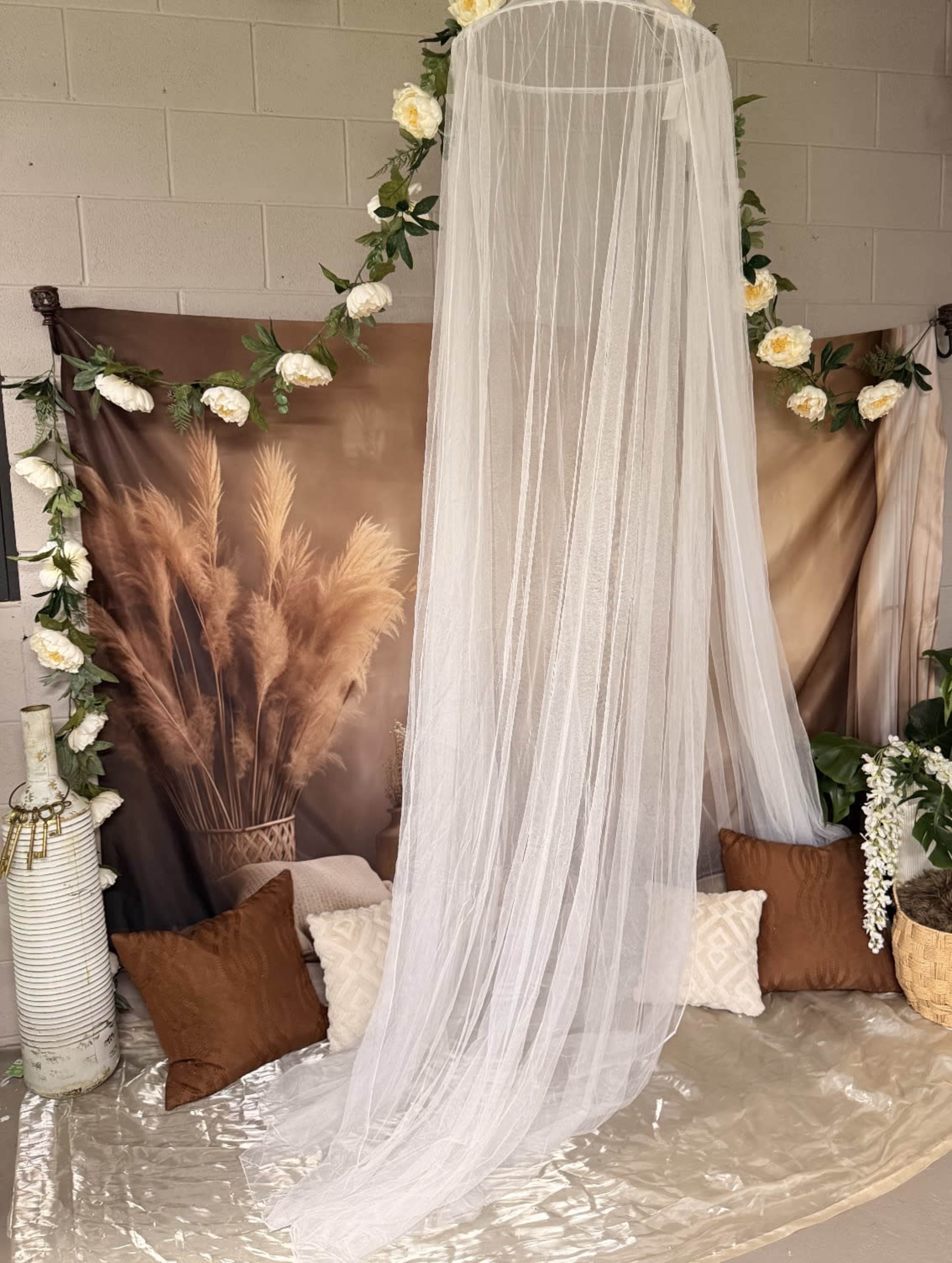 The scene features a draped sheer fabric hanging elegantly with a backdrop of dried grasses and floral decorations, accompanied by cushions arranged on the floor.