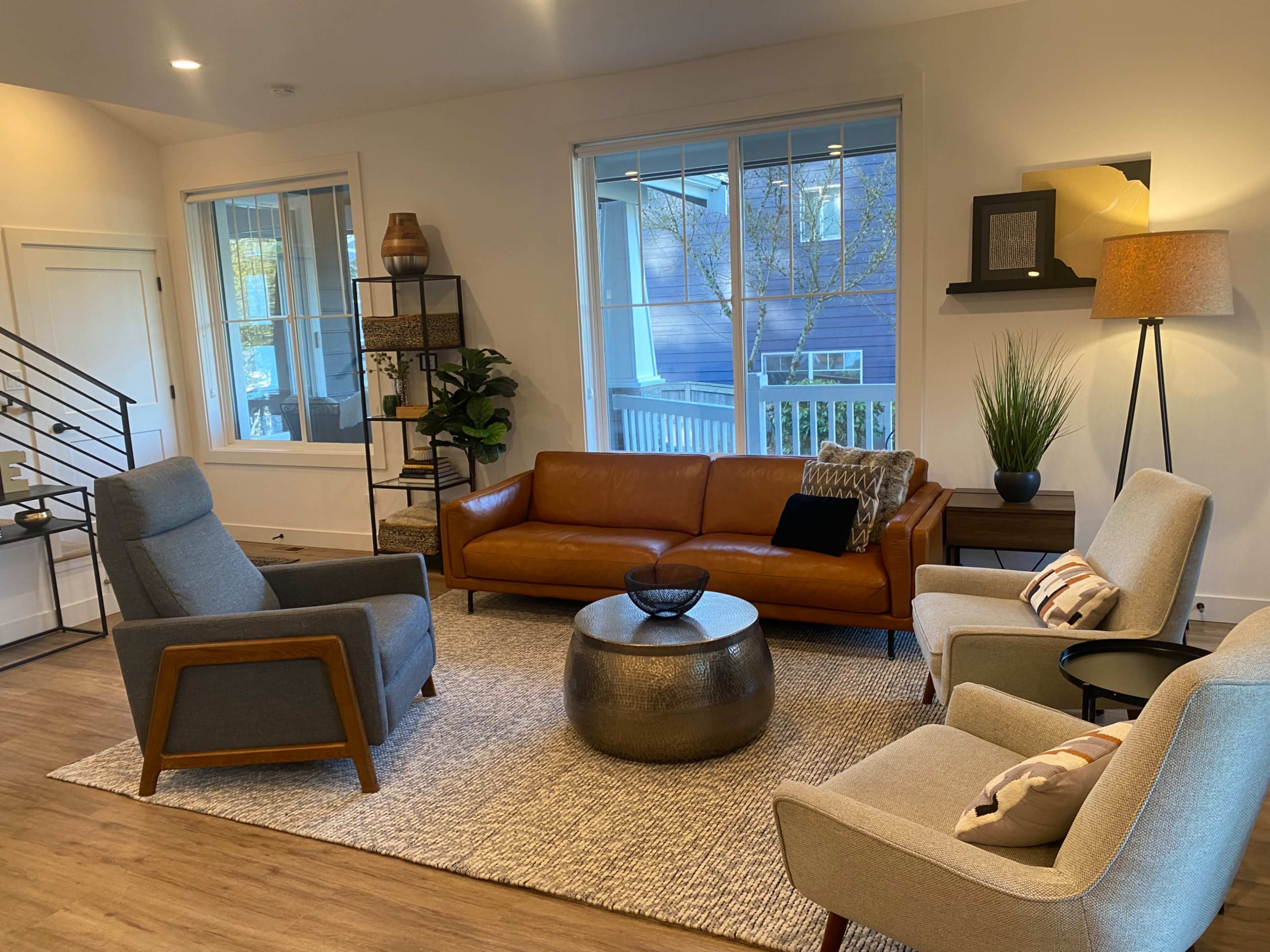 The living room features a brown leather sofa, two gray armchairs, a round metallic coffee table, and large windows showcasing outdoor greenery.