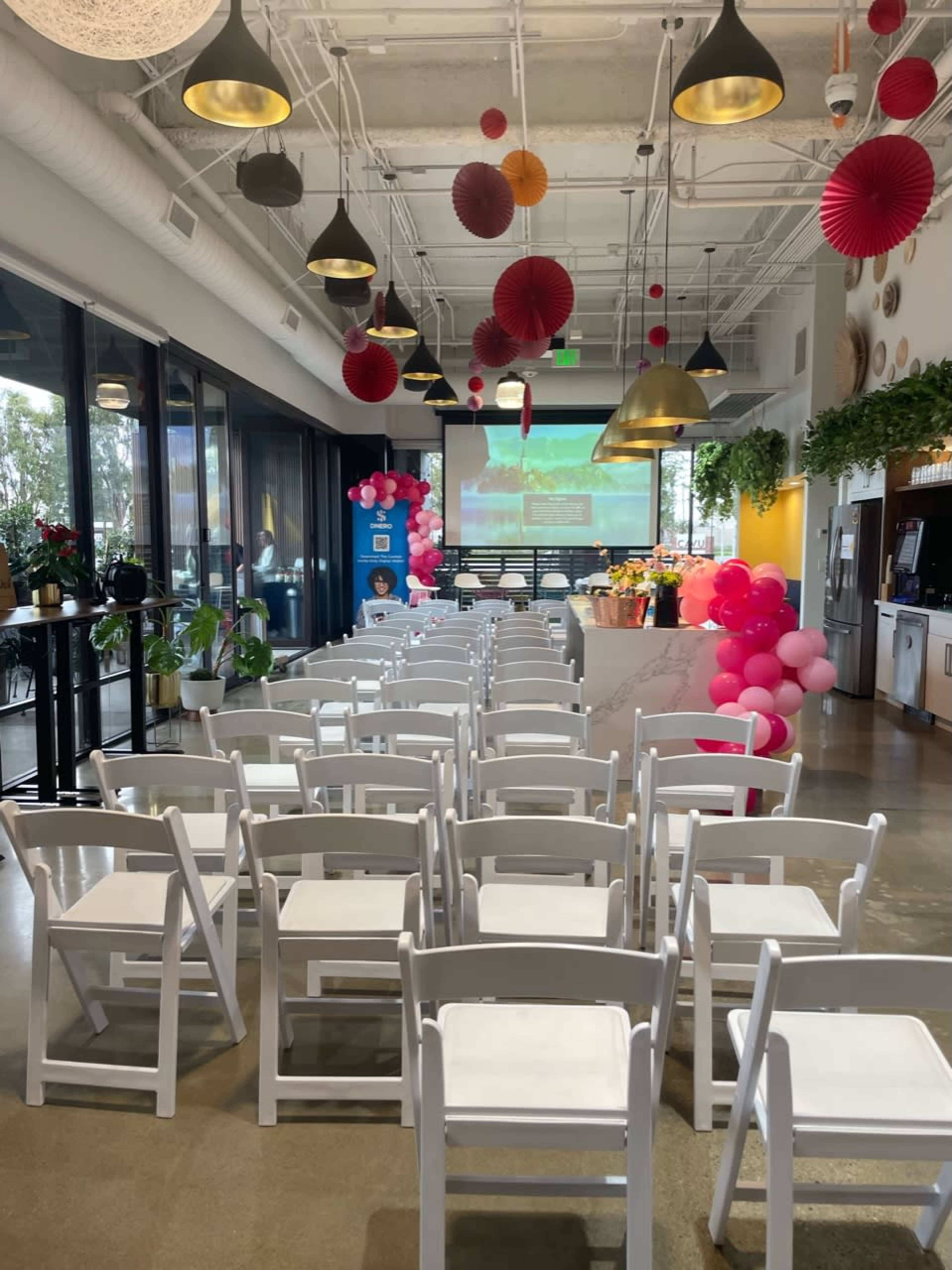 The scene shows a modern event space set up with rows of white chairs facing a screen, decorated with pink balloons and paper lanterns.