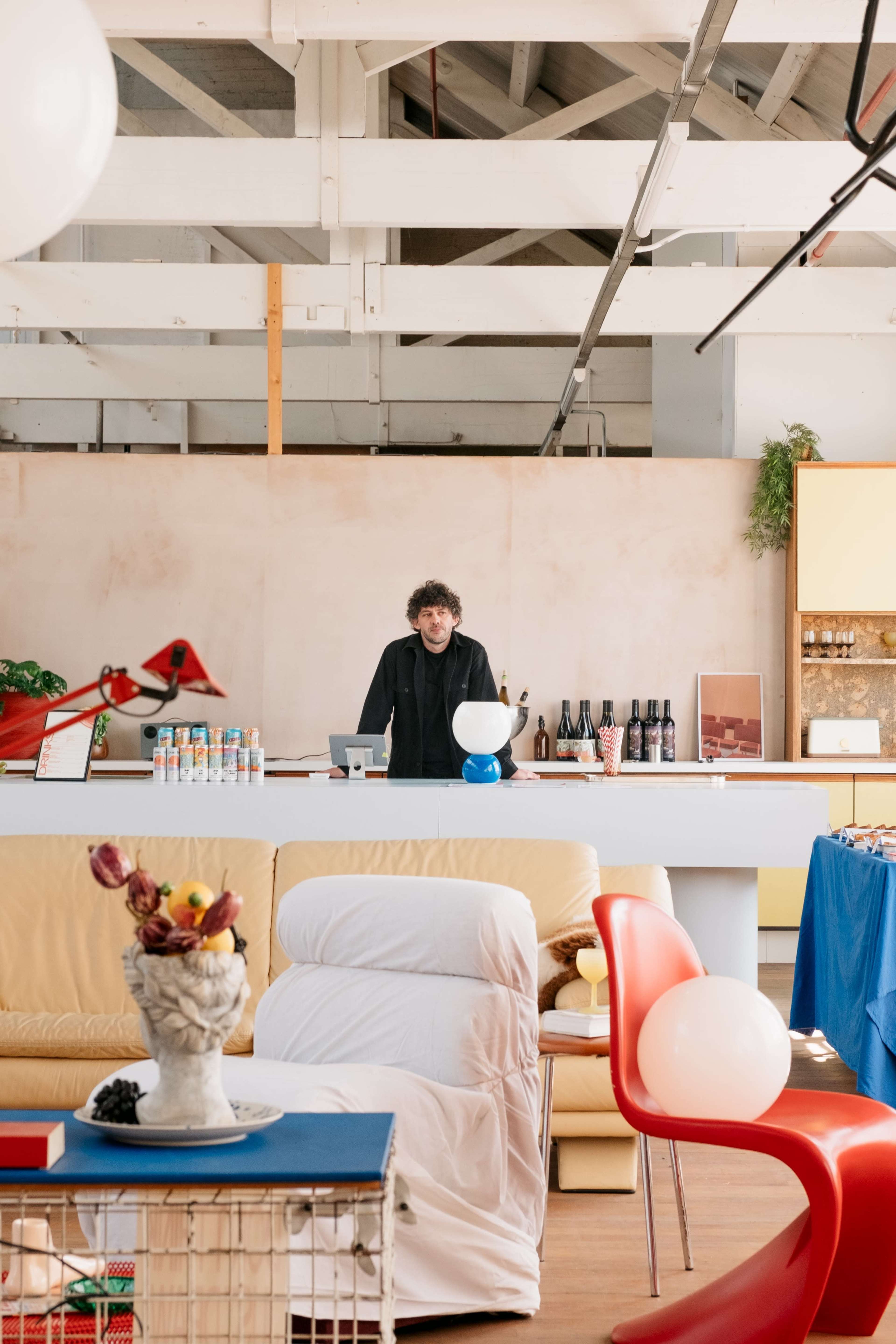 A man stands behind a counter in a modern, stylish interior filled with colorful furniture and decor.