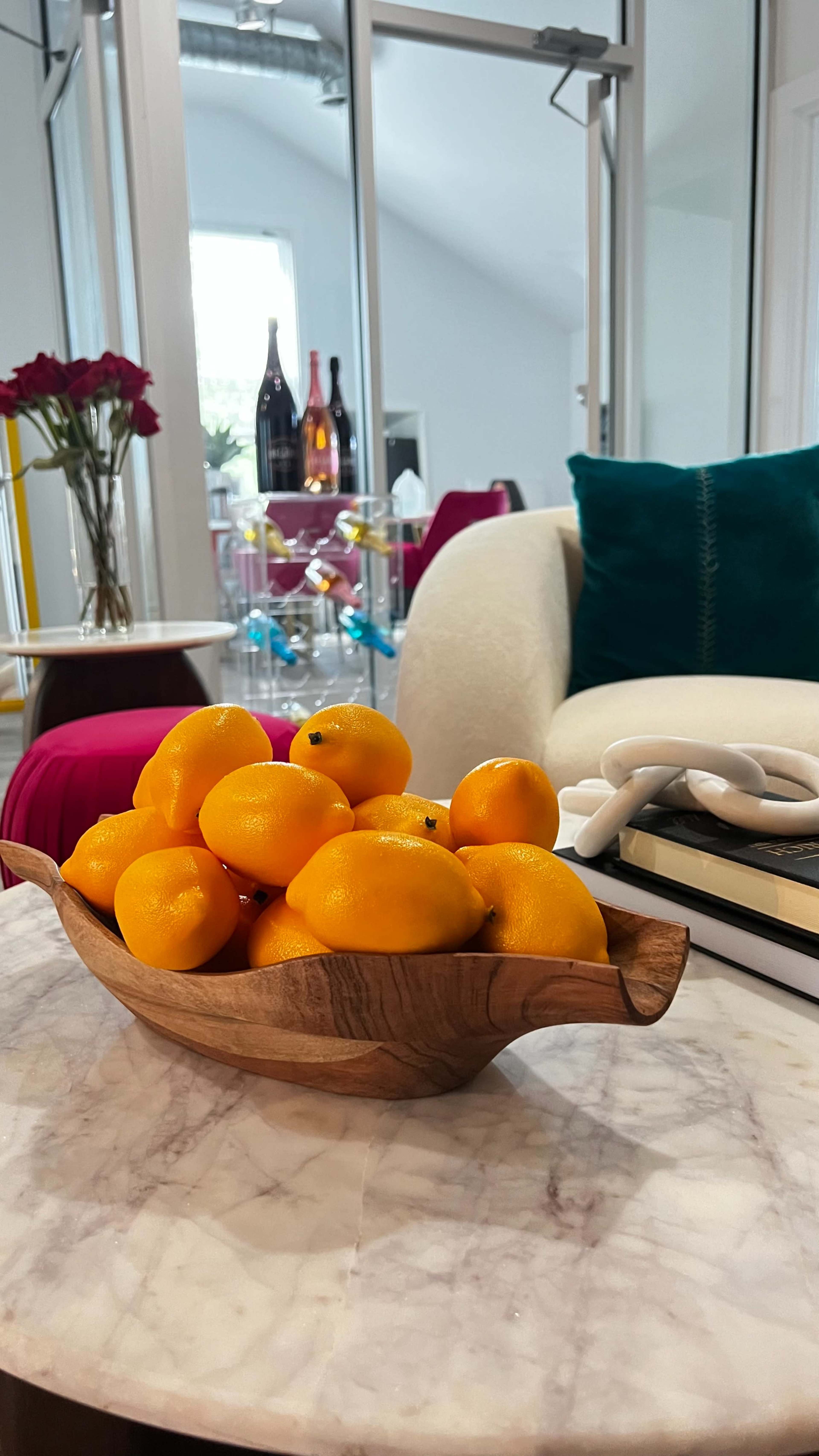 A wooden bowl filled with bright oranges sits on a marble table in a living room with decorative elements in the background.