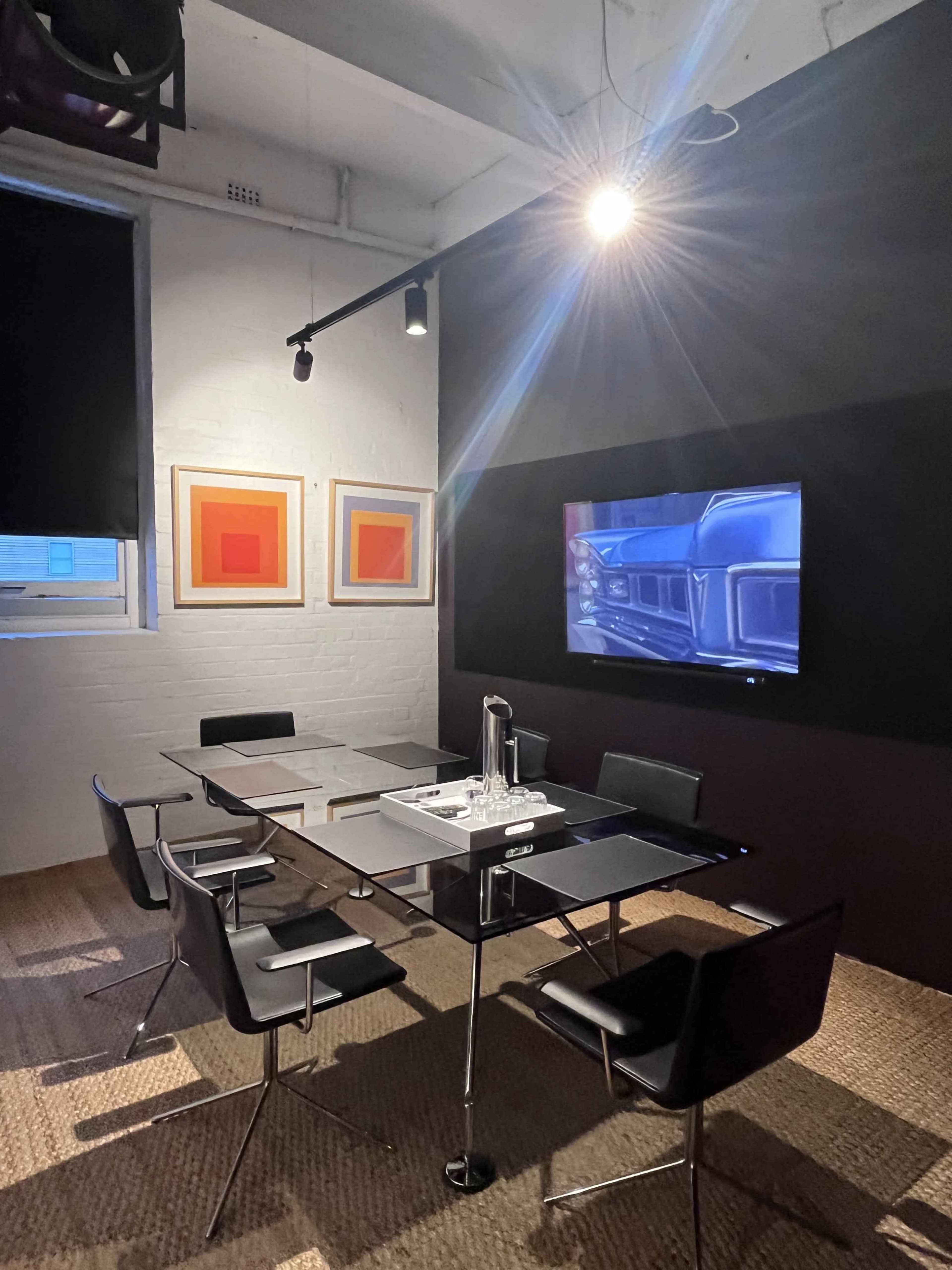 A modern meeting room features a glass-topped table surrounded by black chairs, with framed artwork on the walls and a screen displaying an image.