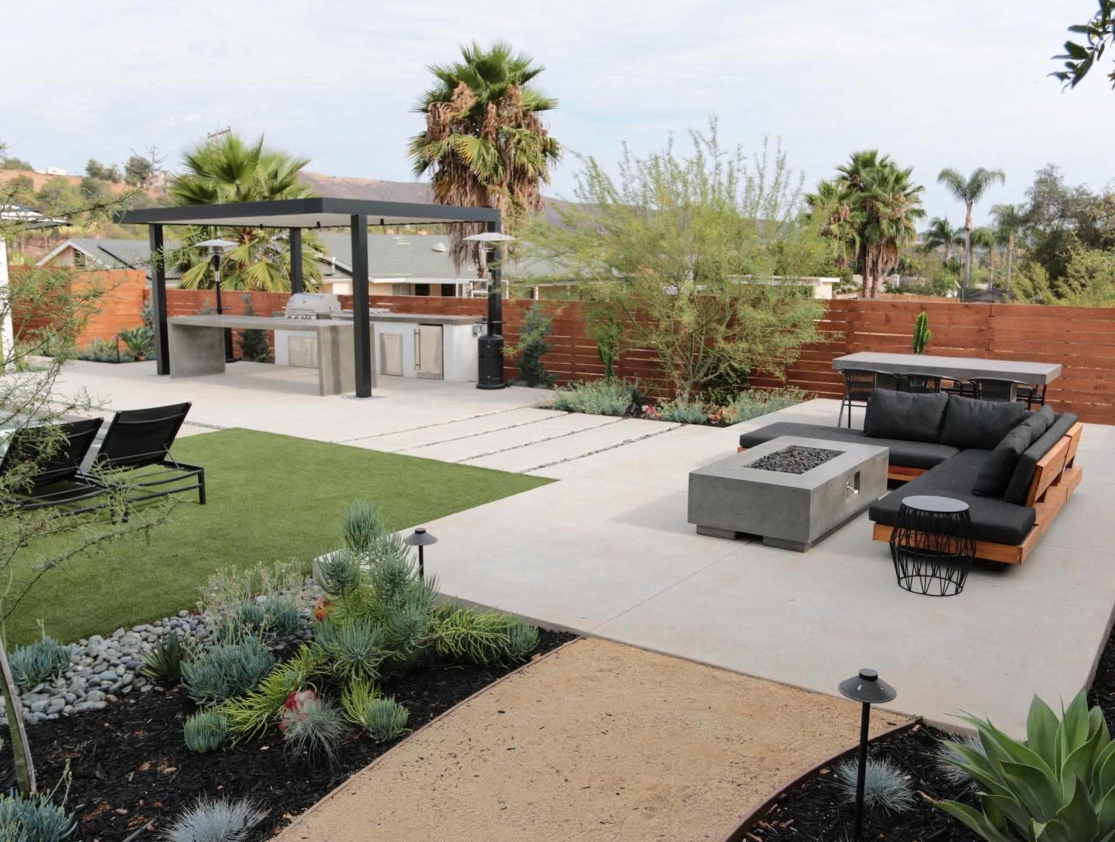 A modern outdoor patio featuring a seating area with a fire pit, an outdoor kitchen, and landscaped greenery with palm trees.