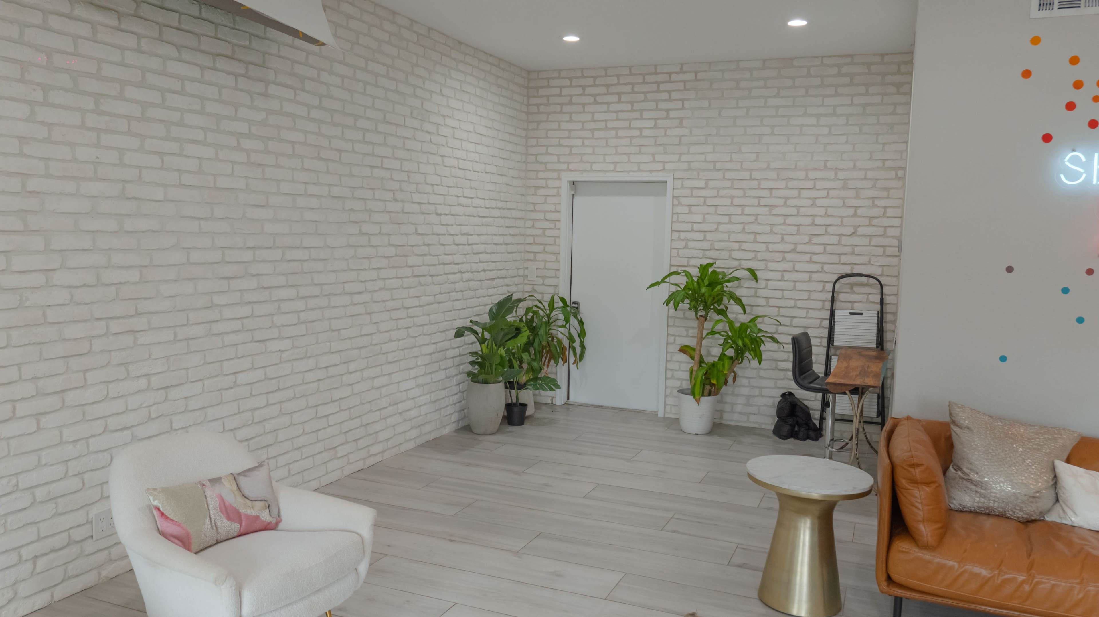 A bright, modern interior space with a white brick wall, potted plants, a small round table, a white chair, and a wooden desk with a chair against the wall.
