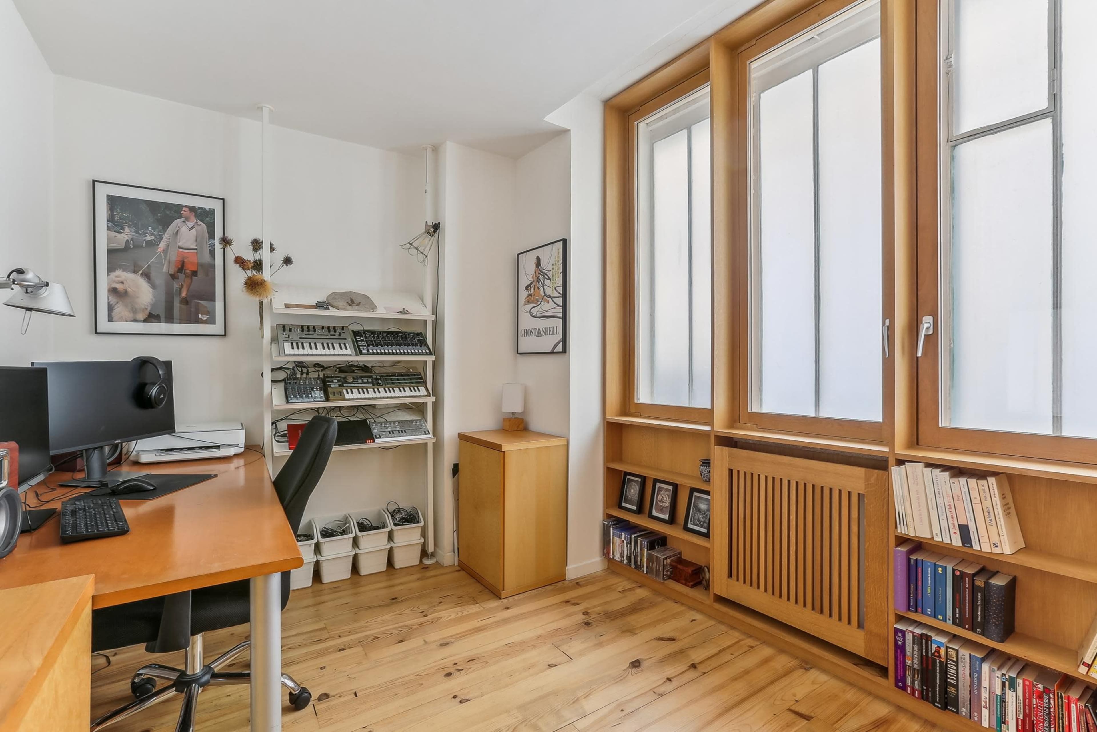 The image shows a small, well-lit office space with a wooden desk, a computer setup, shelving with books, and large windows.