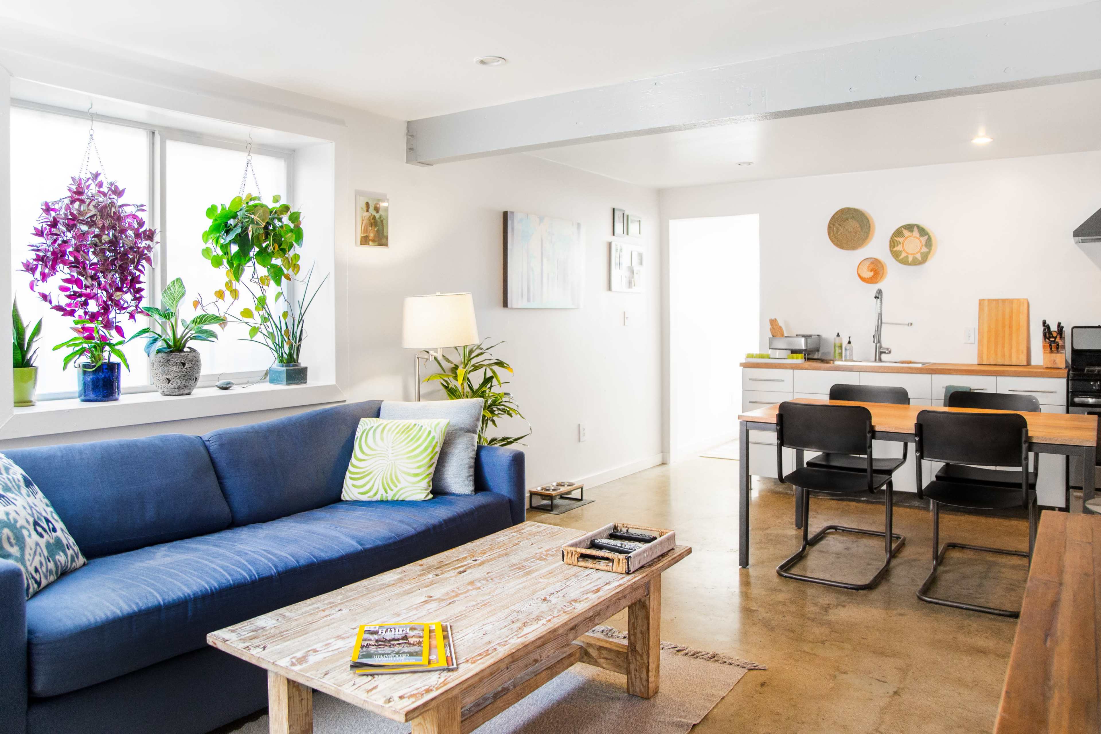 A bright, modern living room with a blue couch, a wooden coffee table, and a kitchen area in the background featuring a minimalist design.