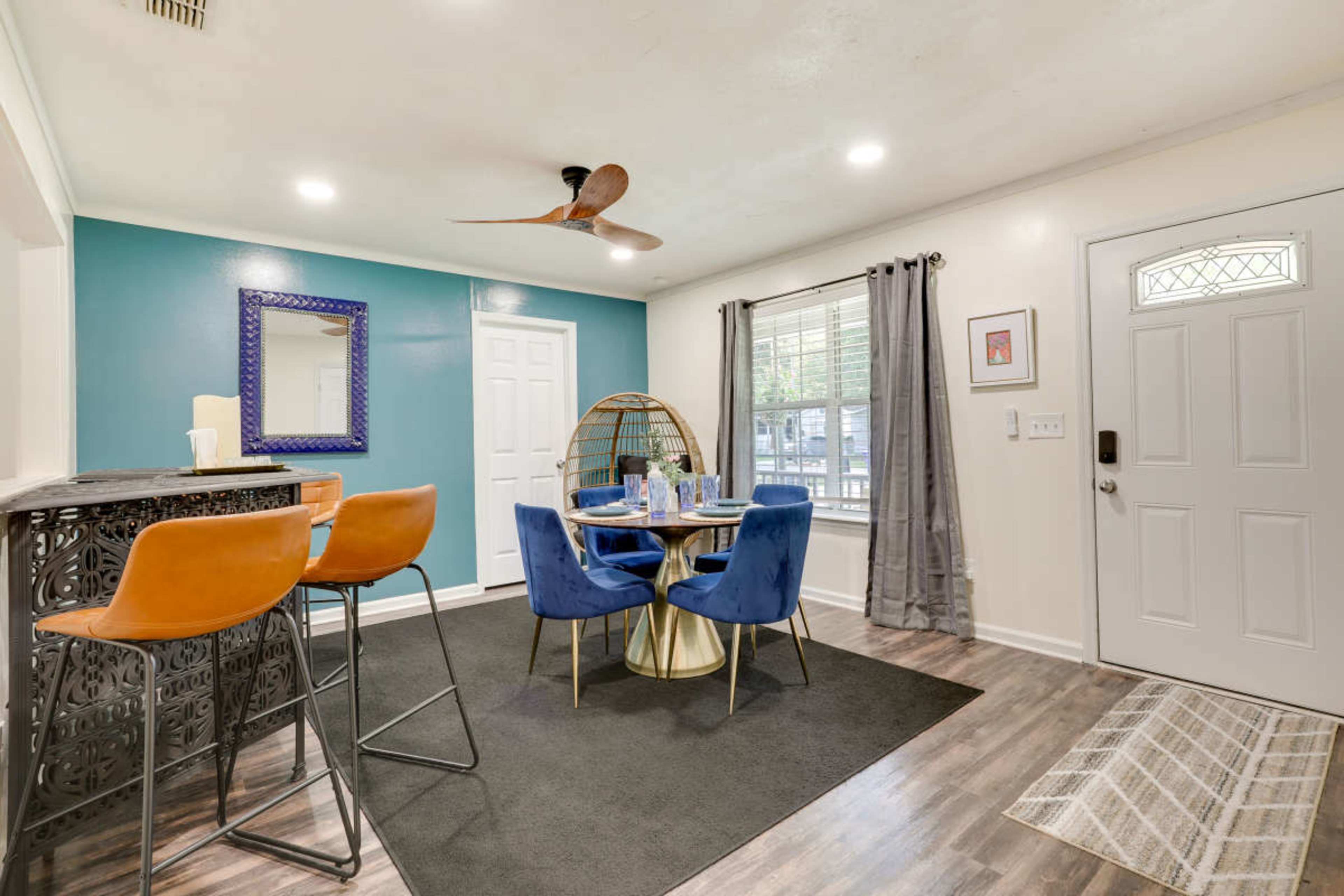 A dining area with a round table set for four, surrounded by blue chairs, and features a modern bar with two stools, a decorative mirror on the wall, and a front door with a window.